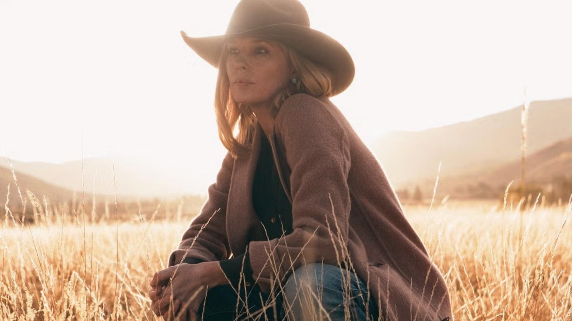 Beth Dutton crouches in the golden field at sunset wearing a wide-brim cowboy hat and long brown wool coat.