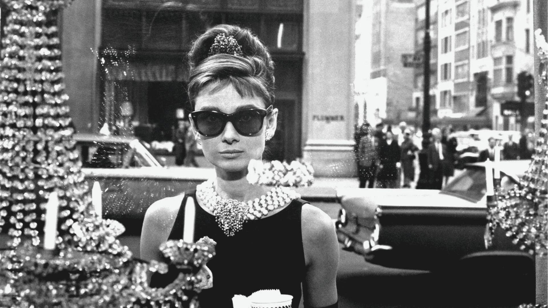 Iconic scene from Breakfast at Tiffany's: Audrey Hepburn as Holly Golightly in a Givenchy little black dress, layered pearl necklace, and oversized sunglasses.