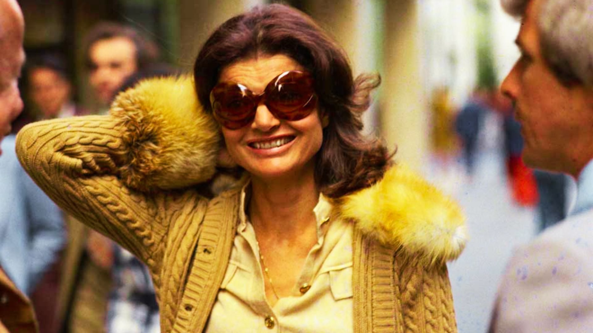 Jackie Kennedy Onassis smiles on a busy street in oversized tortoiseshell sunglasses and a camel cable-knit cardigan.