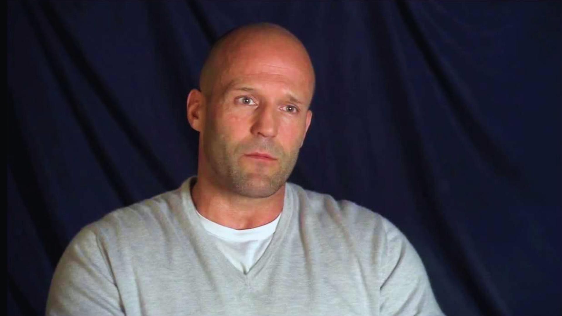 Jason Statham sports a clean shaved head with light stubble beard in a grey sweater, his classic bald style.