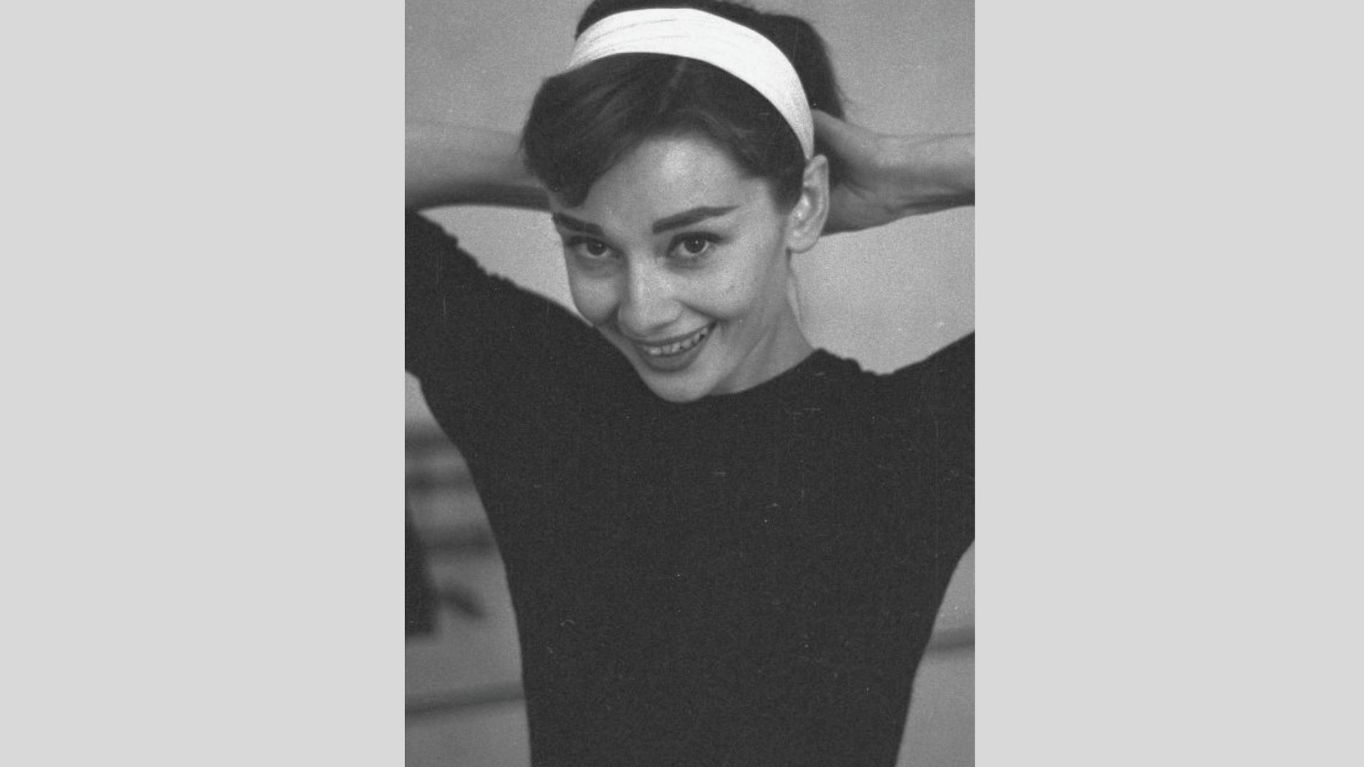 Black-and-white portrait of Audrey Hepburn smiling in a fitted black top and white headband, hands behind her head.