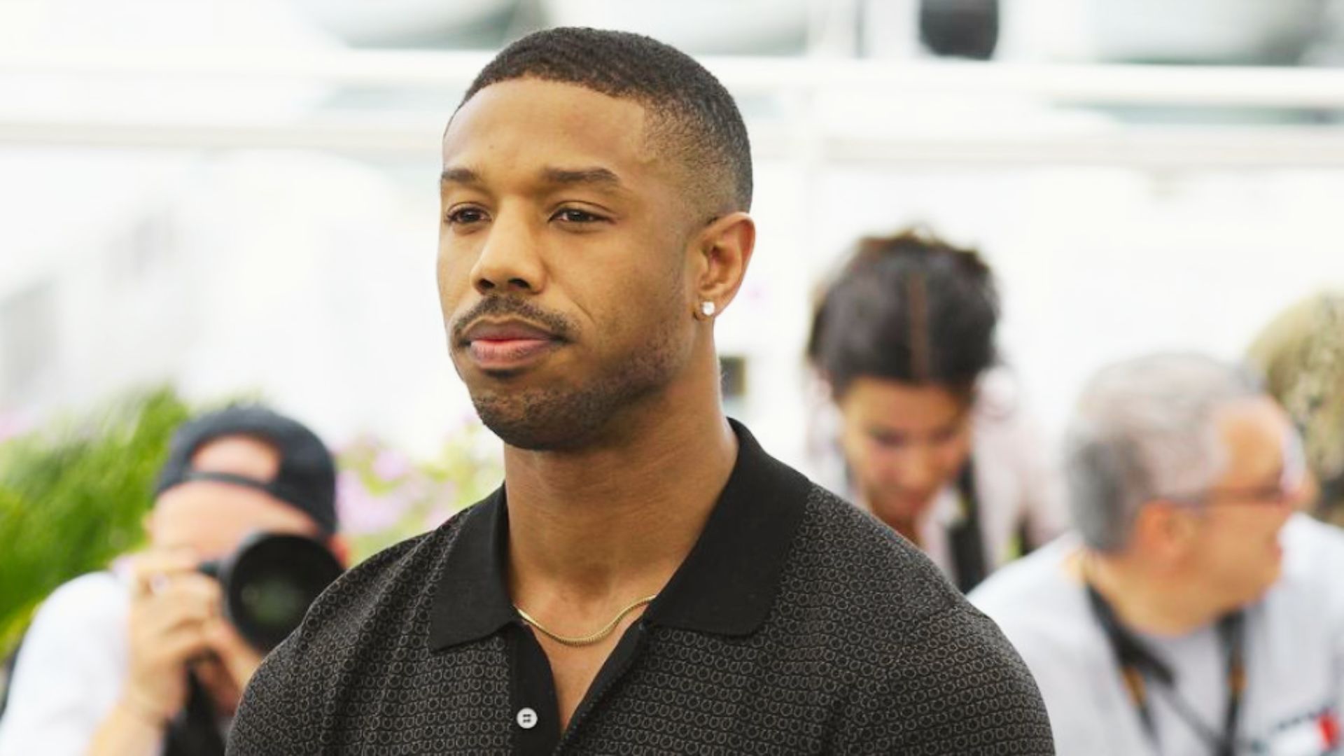 Michael B. Jordan at an outdoor event wearing a black polo, sporting a low fade haircut with a sharp line-up.