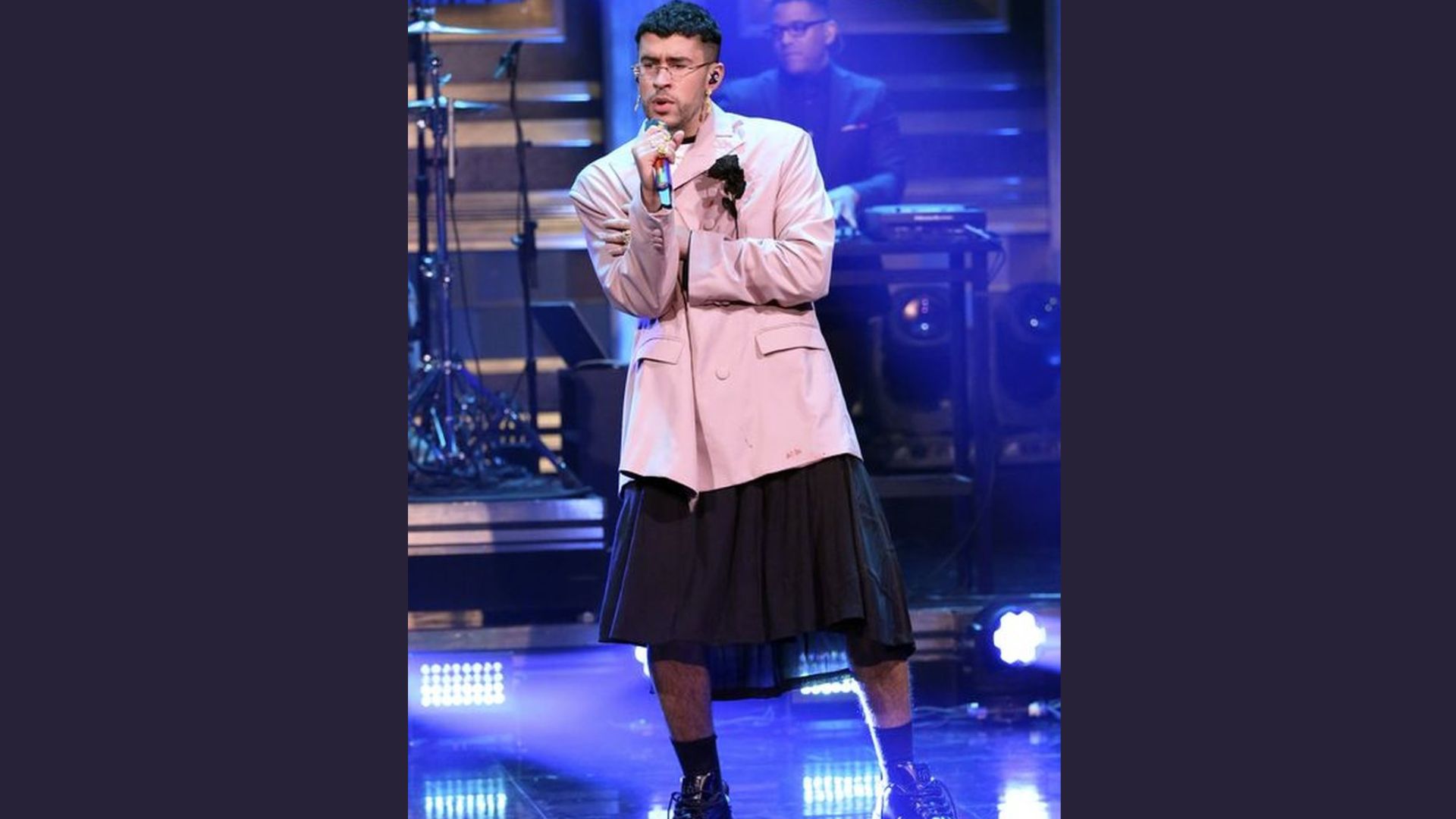 "A performer in a beige blazer and black skirt sings passionately on stage. Behind are band members and blue lighting, creating a lively atmosphere."