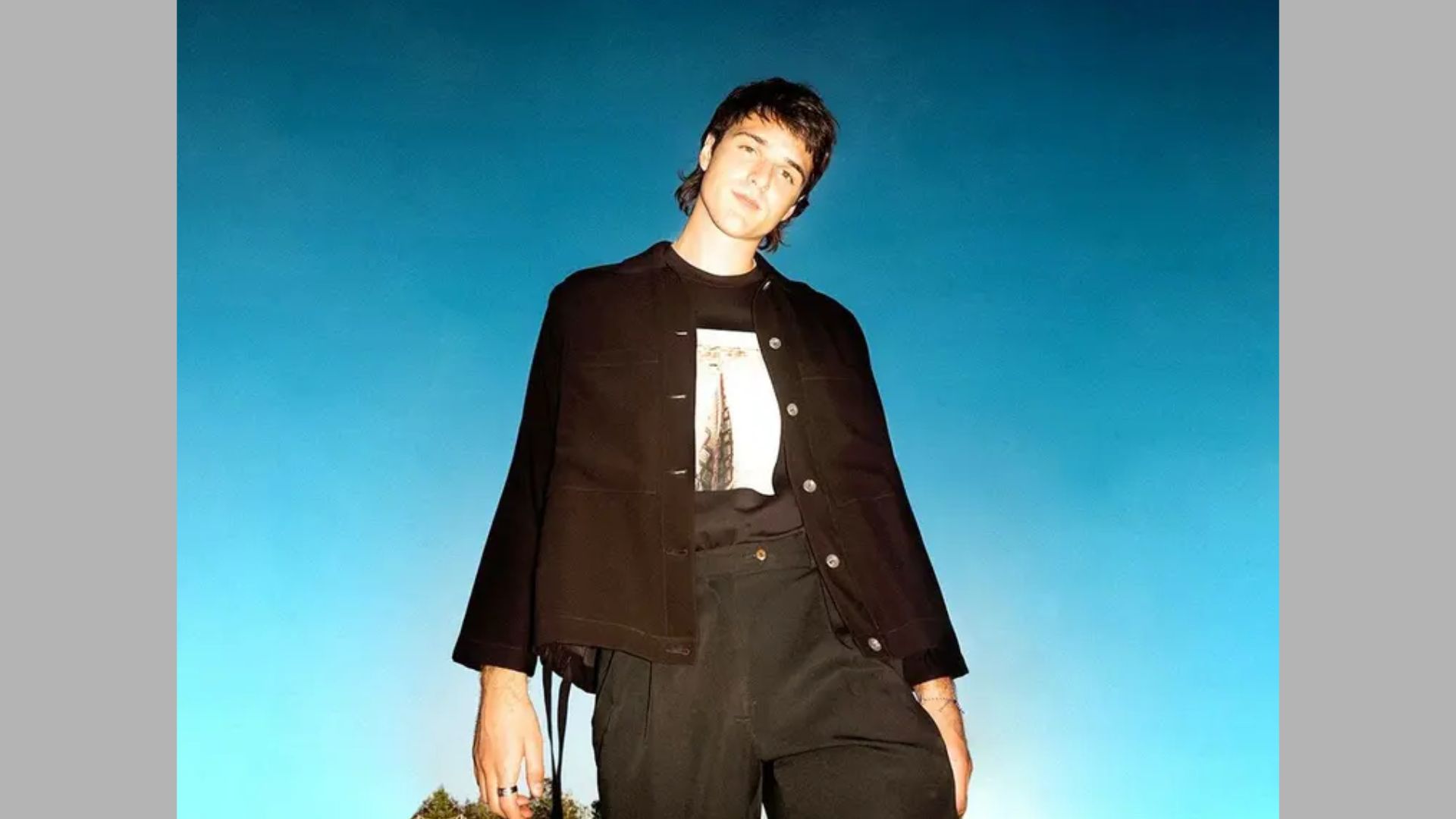 A man in a black jacket and pants stands confidently on a hill, overlooking the landscape below.