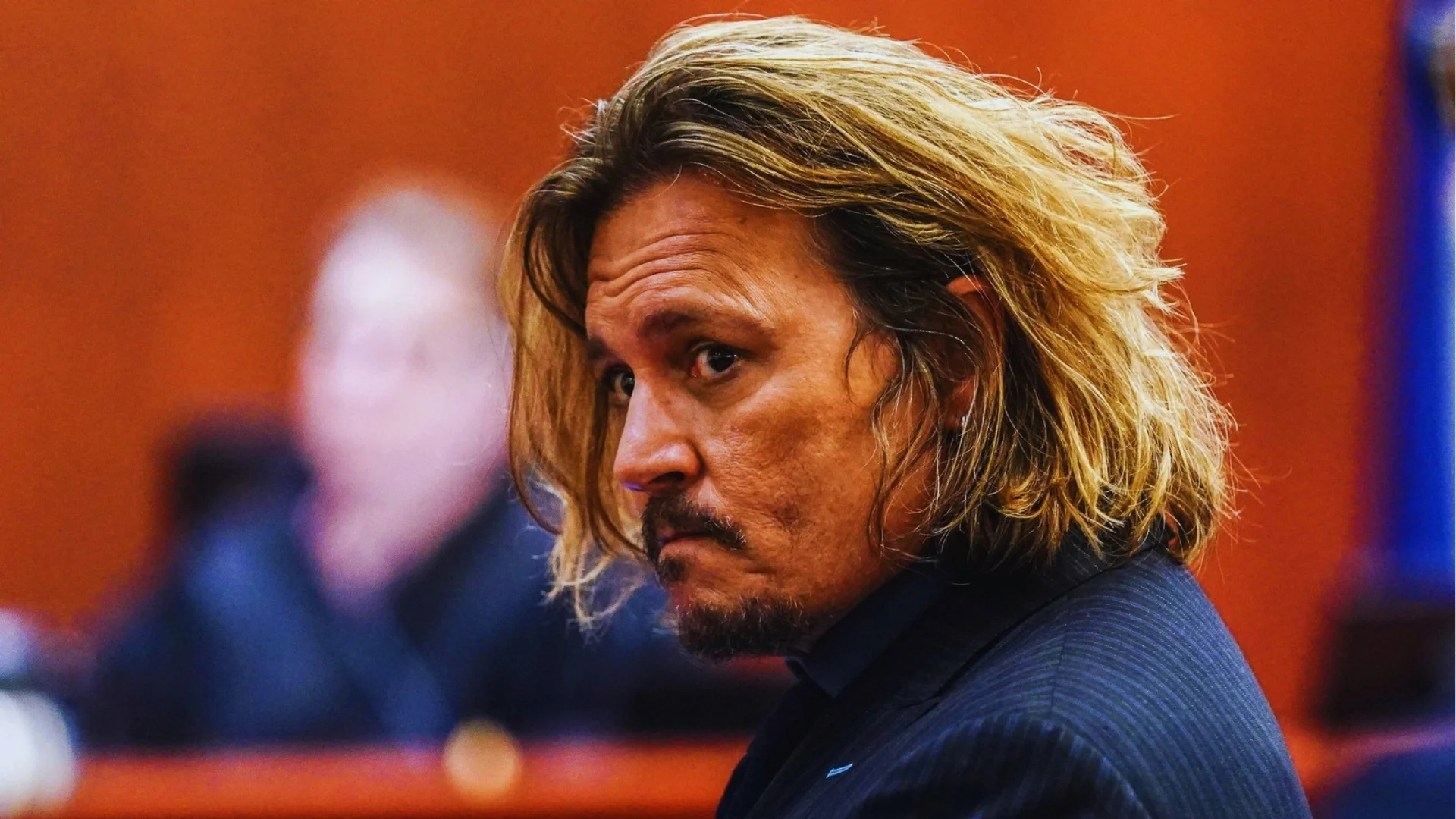 Johnny Depp in the courtroom with long, tousled, layered blond hair and goatee wearing a dark suit.