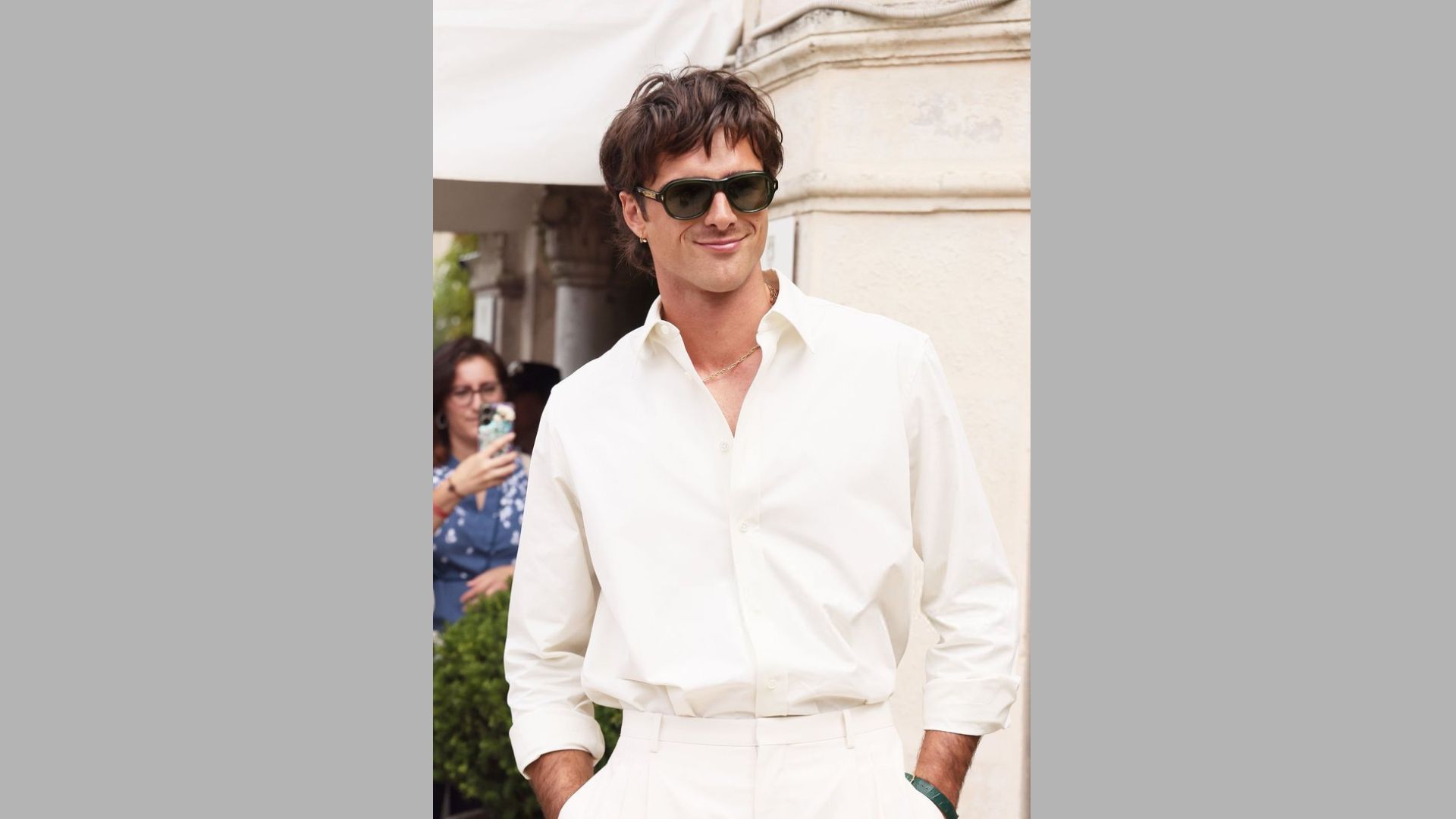 A man wearing a white shirt and sunglasses stands confidently, showcasing a casual yet stylish appearance.
