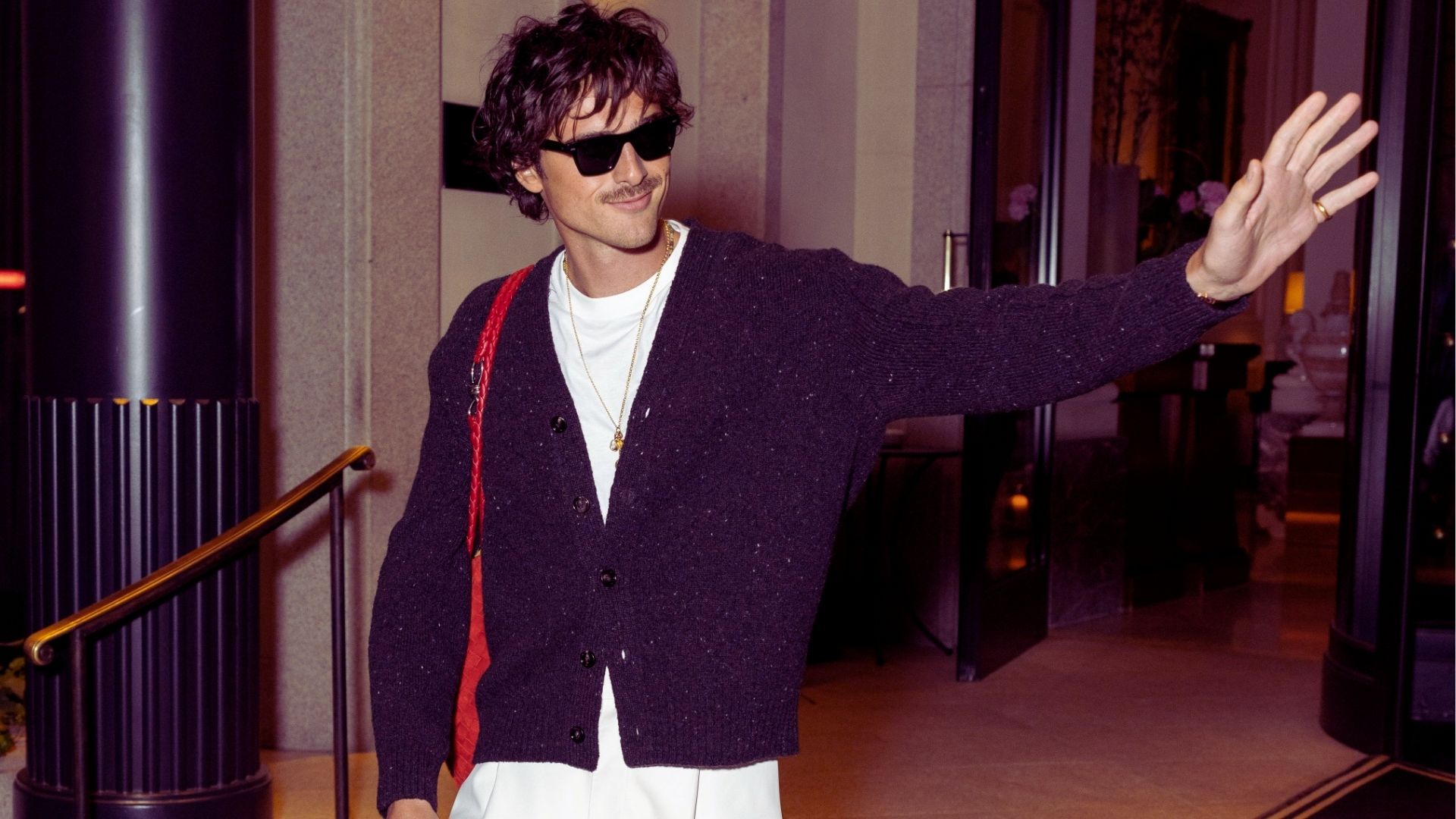A man wearing sunglasses and a sweater waves cheerfully at the camera.