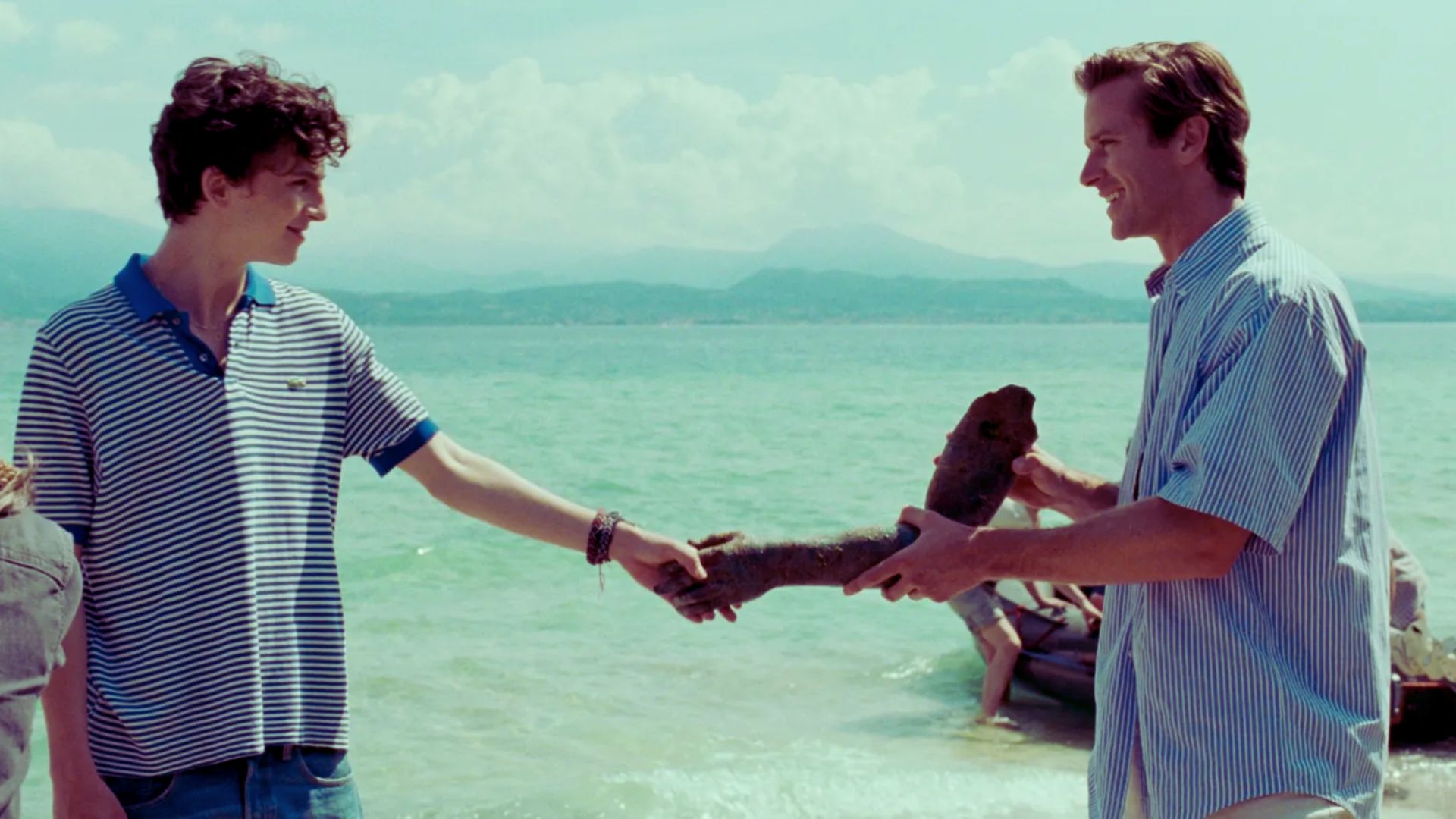 Two men shaking hands on a beach, with a boat visible in the background under a clear blue sky."