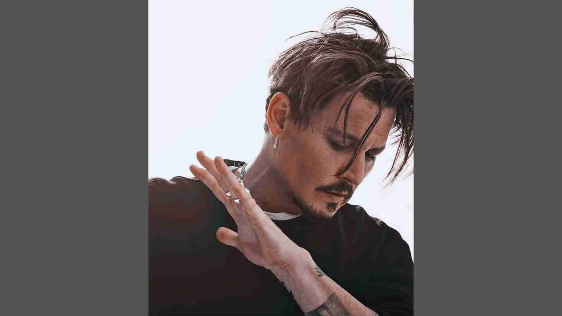 Johnny Depp with messy textured medium-length hair looking down, hand raised, wearing black sweater and rings.