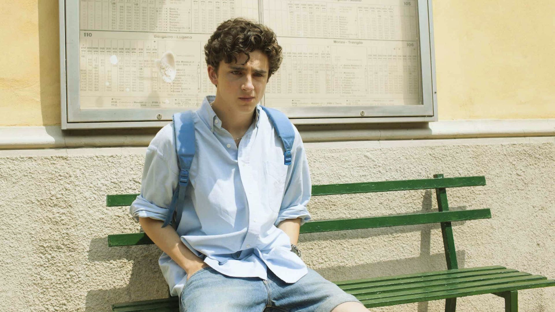 A young man sits on a bench, resting with his backpack beside him in a casual outdoor setting."