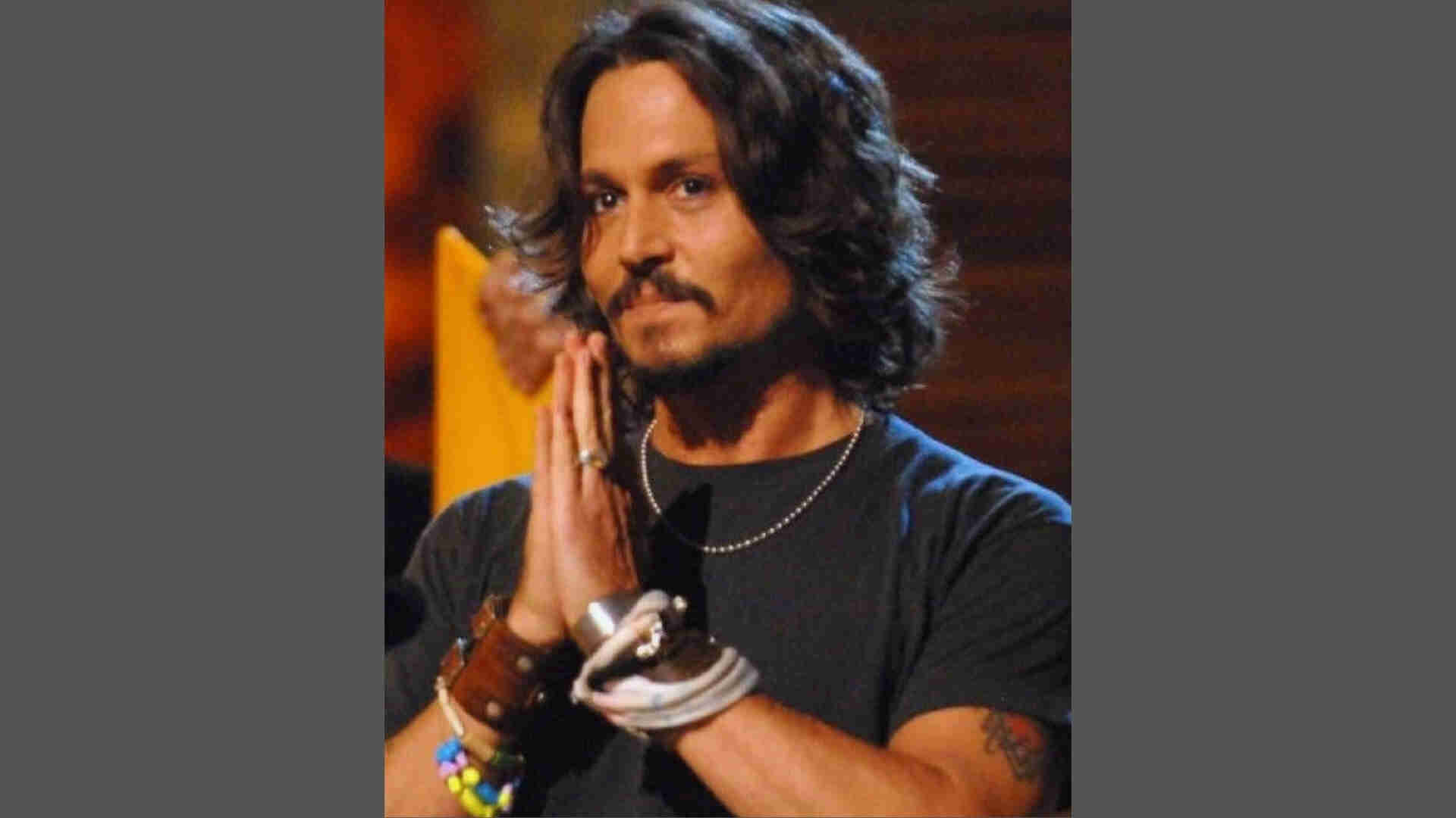 Johnny Depp with long wavy center-parted hair, mustache and goatee, hands together, wearing black t-shirt and bracelets.