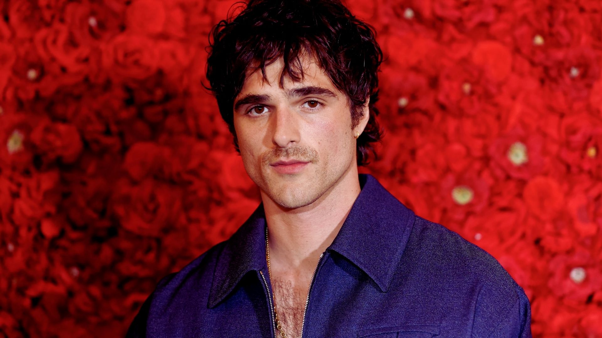 A man with black hair wearing a blue shirt stands confidently, smiling at the camera.