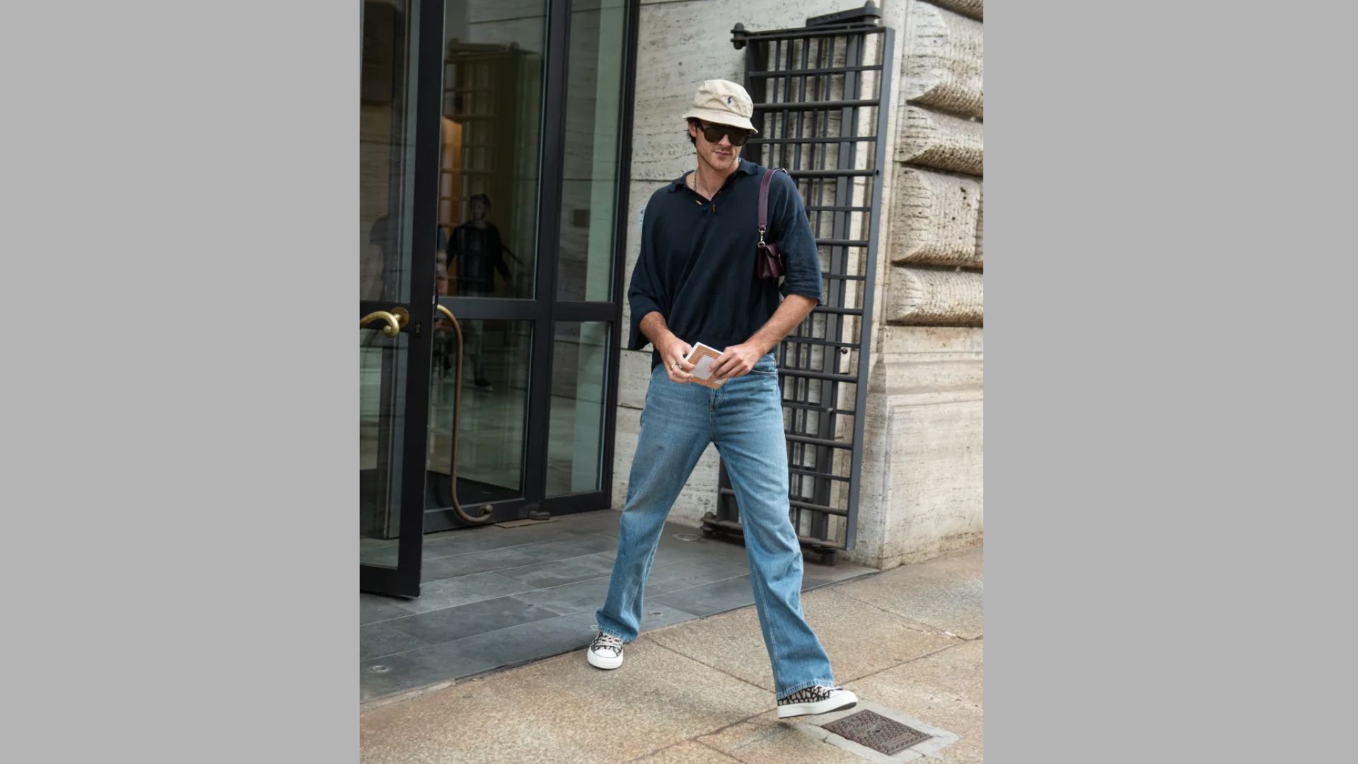A man wearing jeans and a hat walks down a city street, surrounded by buildings and pedestrians.