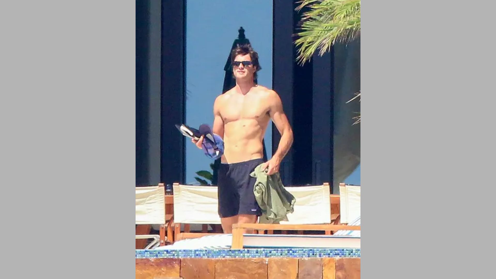 A shirtless man stands by a pool, holding a water bottle, enjoying a sunny day outdoors.