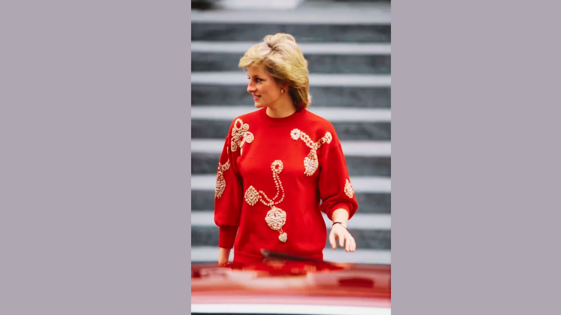 Princess Diana smiles beside a red car in a bold red sweater featuring white embroidered jewelry motifs.