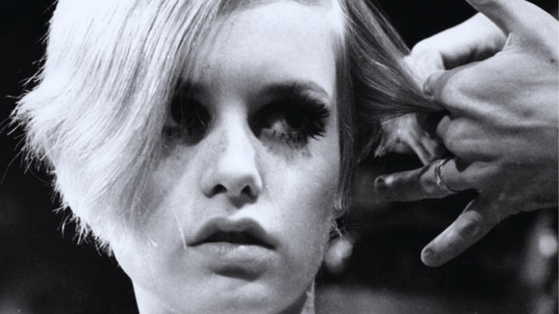 Black and white photo of young Twiggy getting her famous blonde crop cut, deep side part and heavy lashes.
