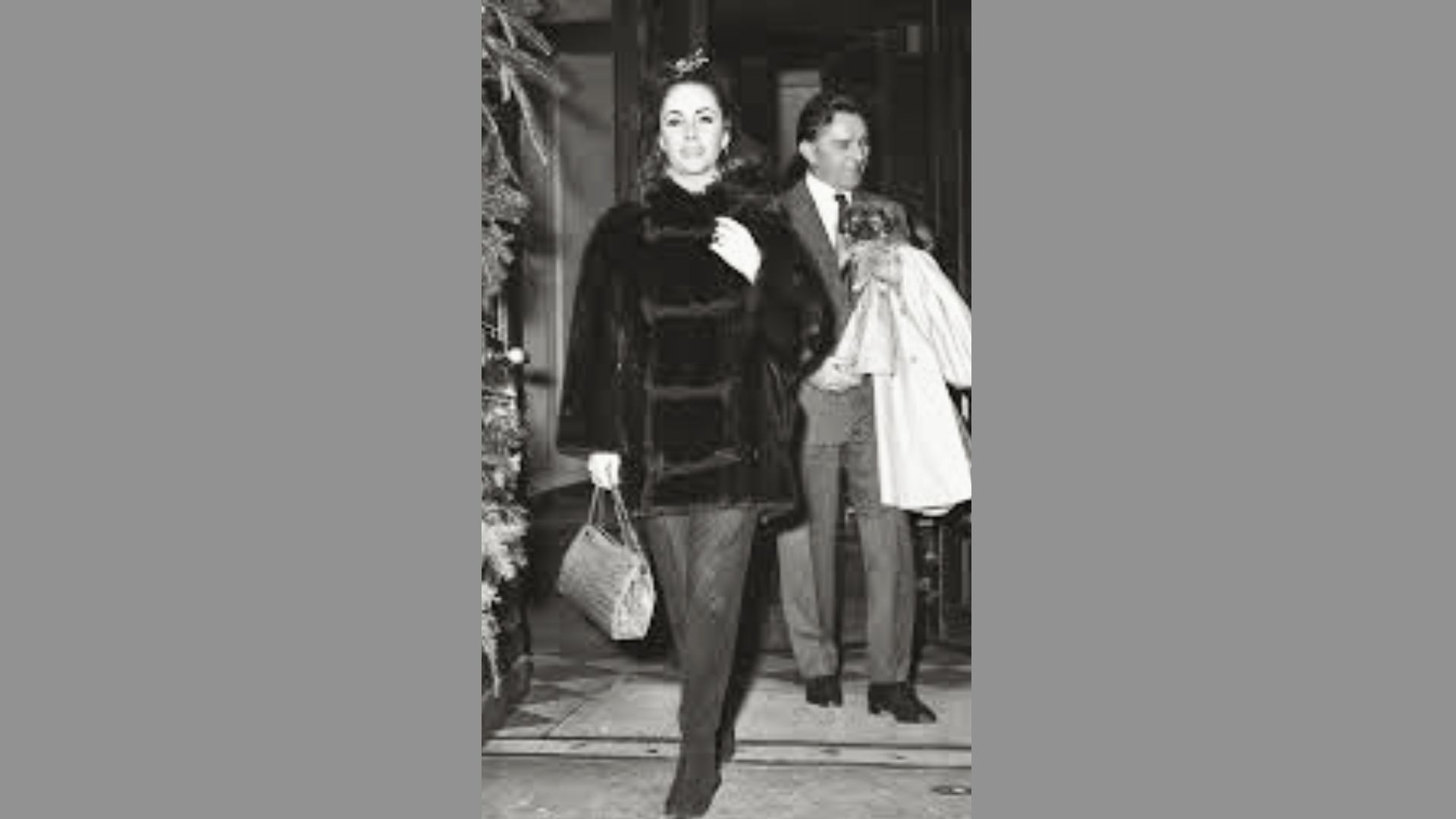 Elizabeth Taylor exits a building in a luxurious dark fur coat paired with slim trousers and a structured handbag.
