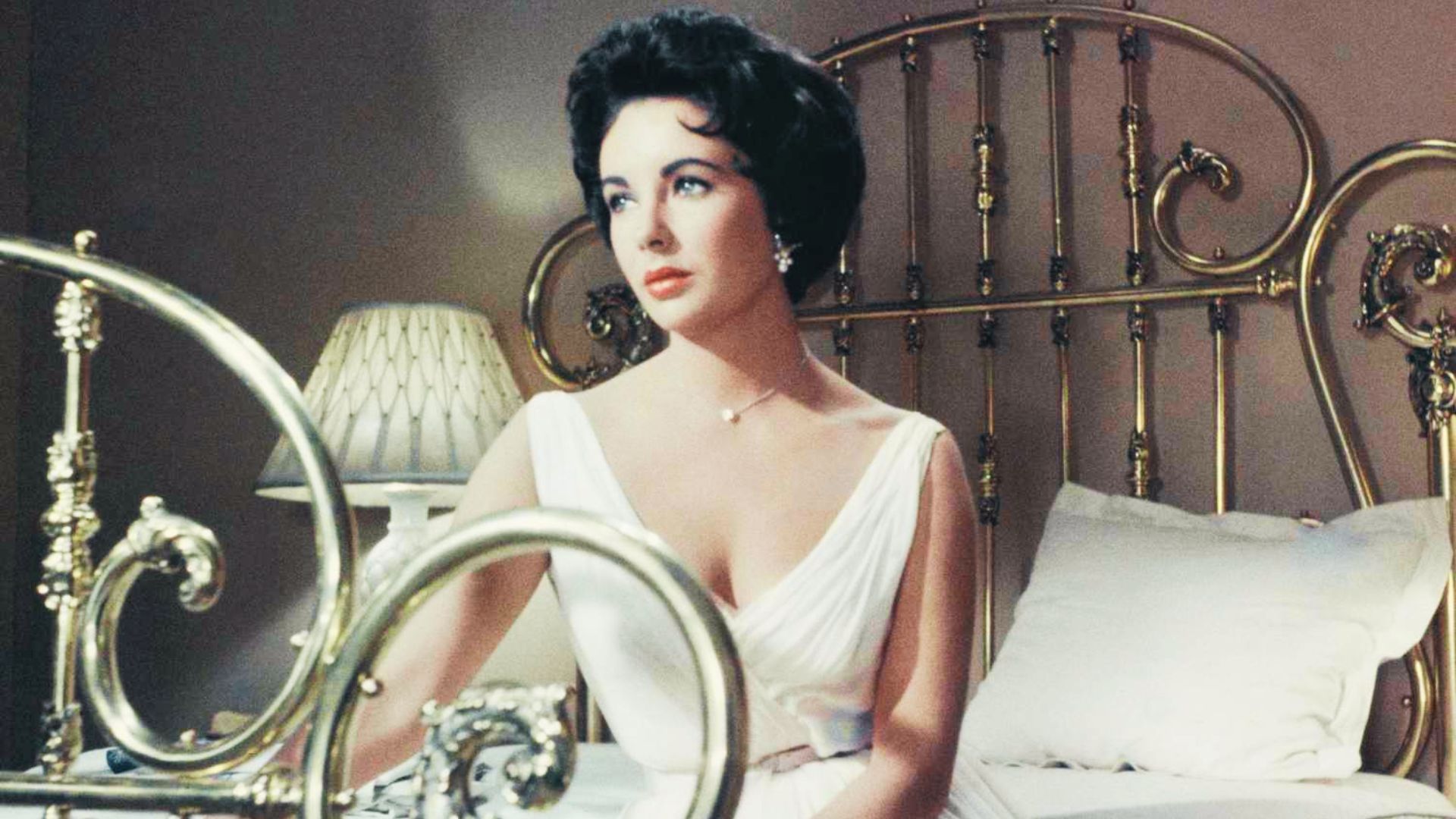 Elizabeth Taylor sits on an ornate brass bed in a draped white V-neck gown from Cat on a Hot Tin Roof.
