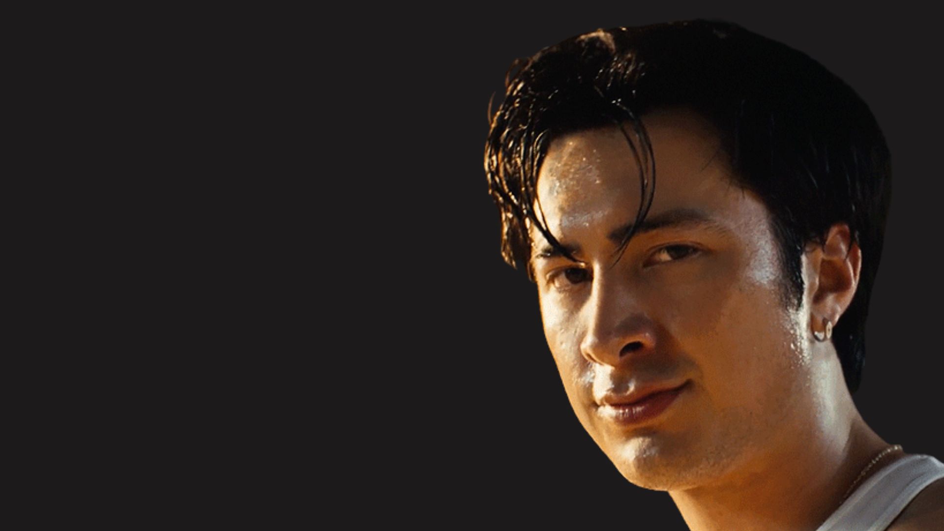 Hudson Williams with slicked back dark hair and a natural smile wearing a white tank top against a dark background.