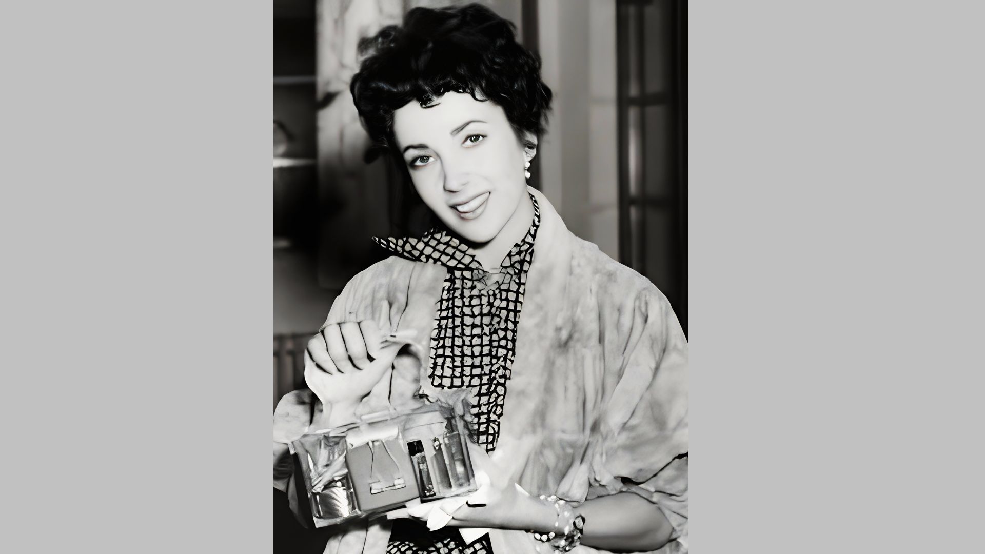 Elizabeth Taylor smiles while holding a clear cosmetic case, wearing a patterned blouse and light jacket.