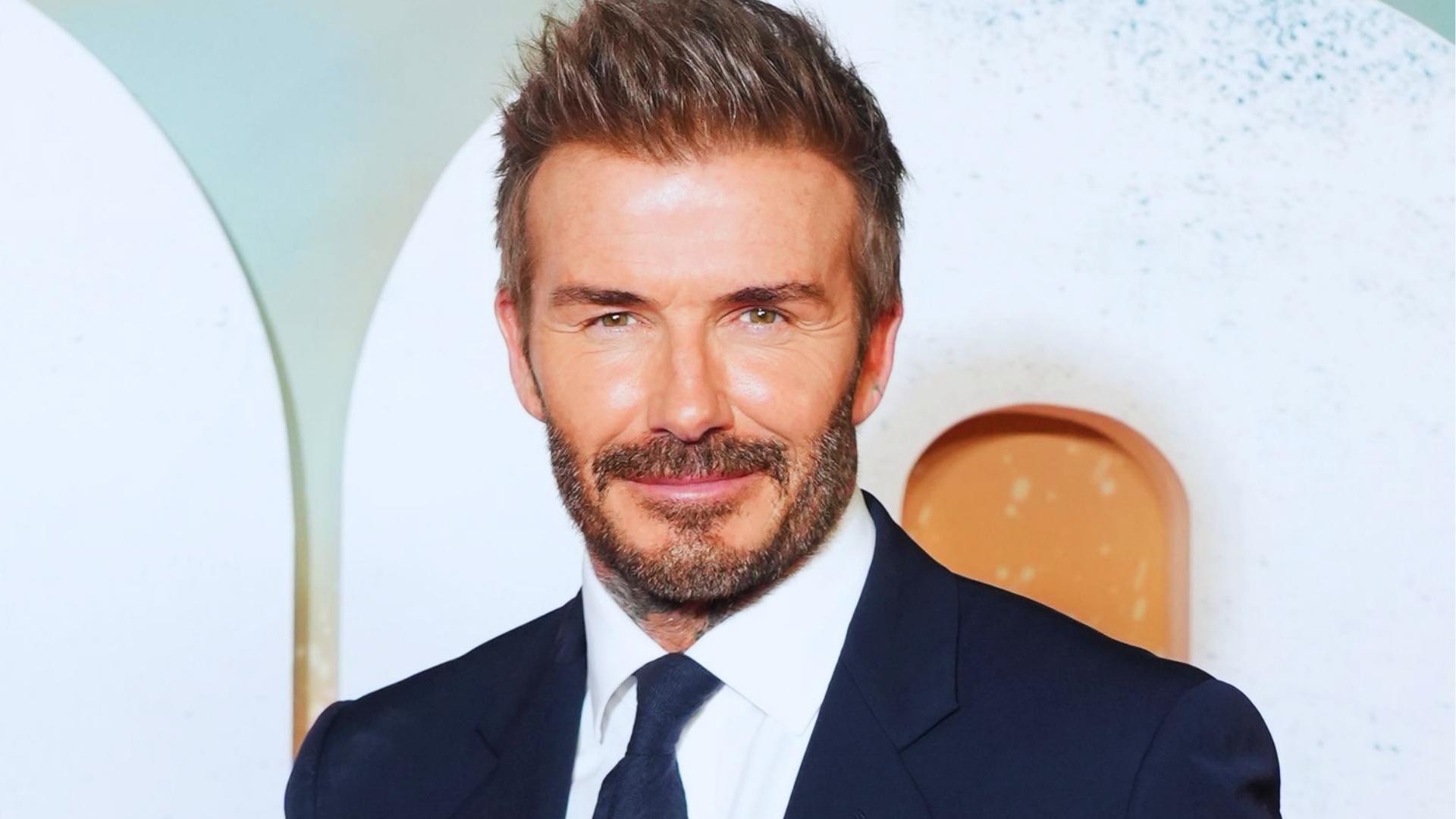 David Beckham smiles at an event with full textured quiff, salt-and-pepper hair, navy suit, showing dense styled result.