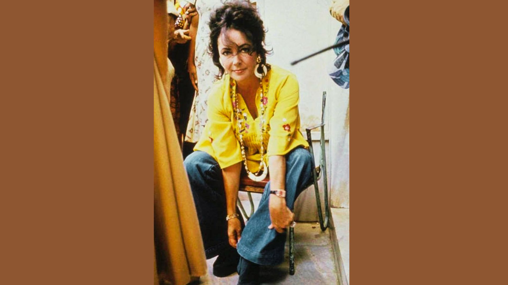 Elizabeth Taylor smiles in a yellow peasant blouse and denim jeans, accessorized with bold jewelry.