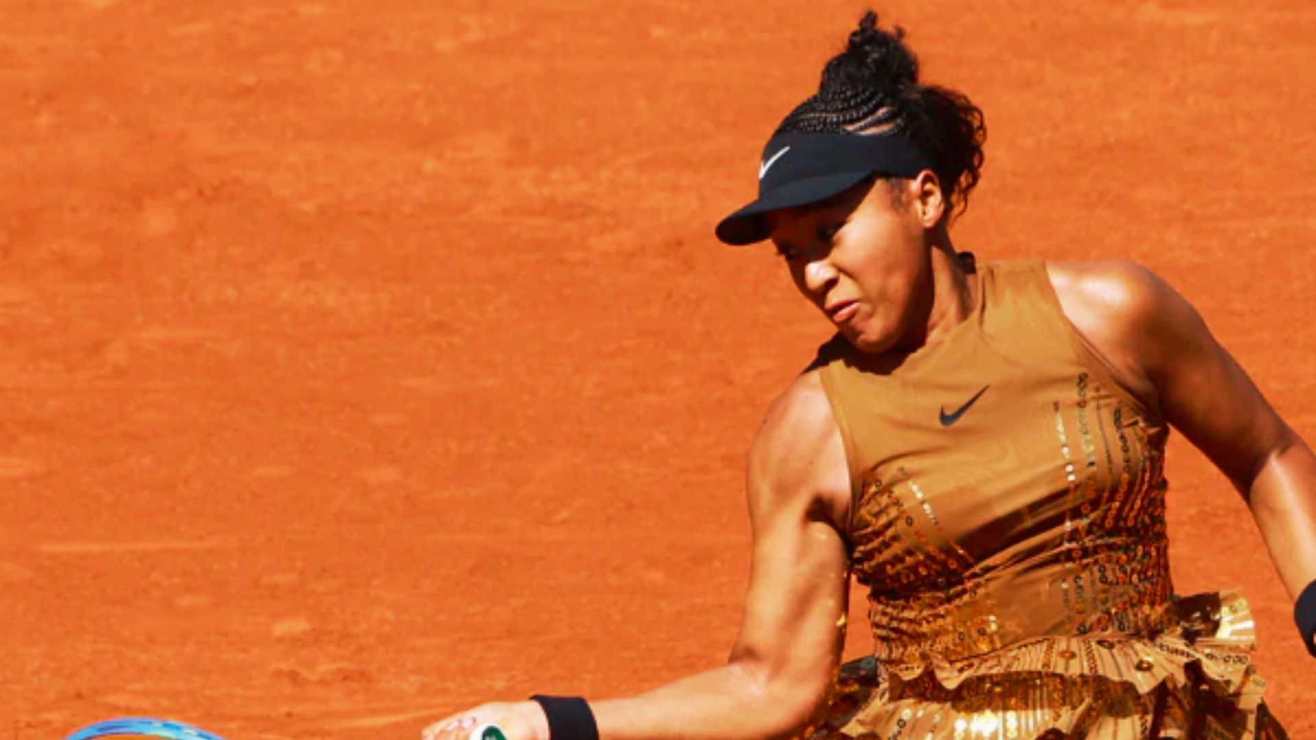 Naomi Osaka in a metallic gold Nike dress with sequin detailing and ruffled hem, mid-swing on a clay court.