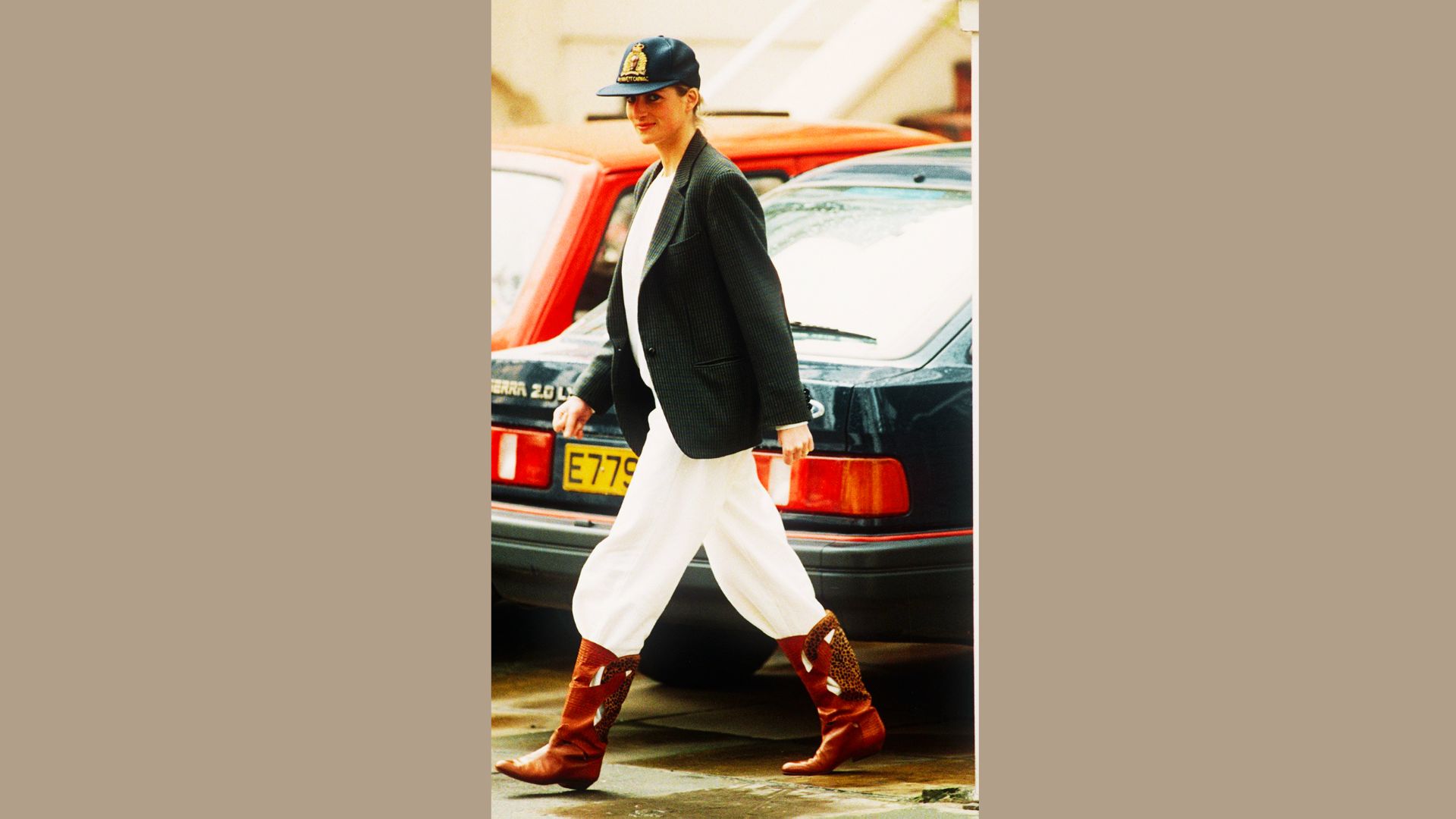 Princess Diana walks city streets in a navy baseball cap, oversized blazer, and white trousers.