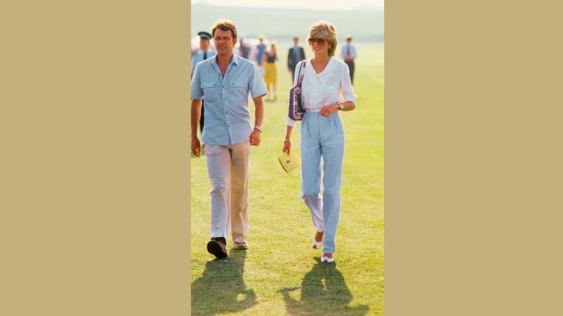 Princess Diana walks across a grassy field wearing a white tie-neck blouse and high-waisted light blue striped trousers.