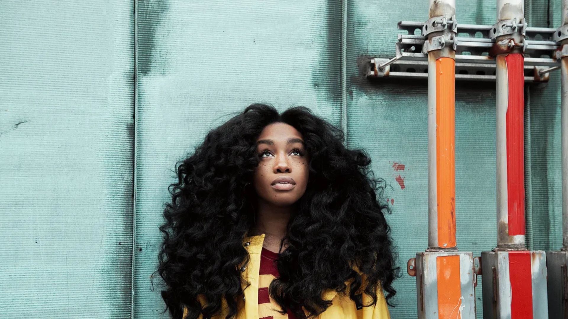 SZA with voluminous afro hair stands confidently in front of a textured wall, showcasing her natural curls.