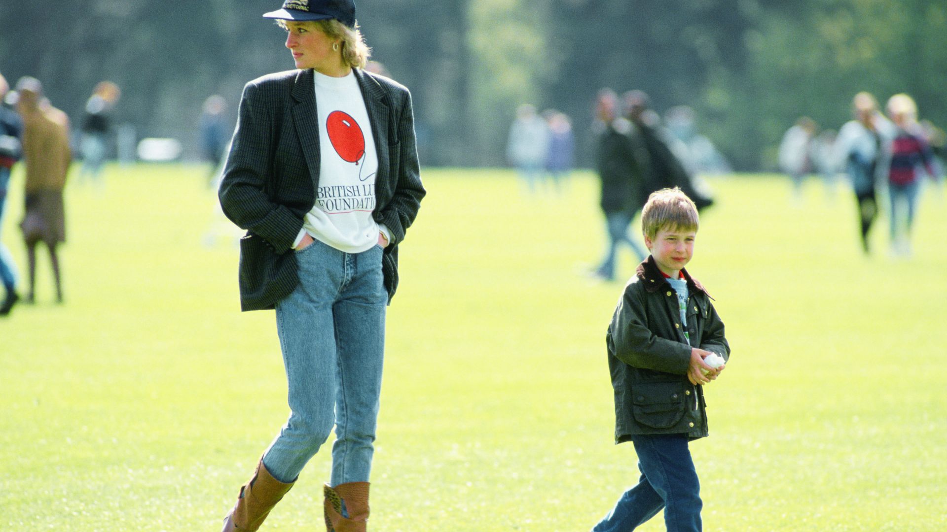 Princess Diana stands on a field with a young Prince Harry, wearing an oversized pinstripe blazer over a charity graphic tee.