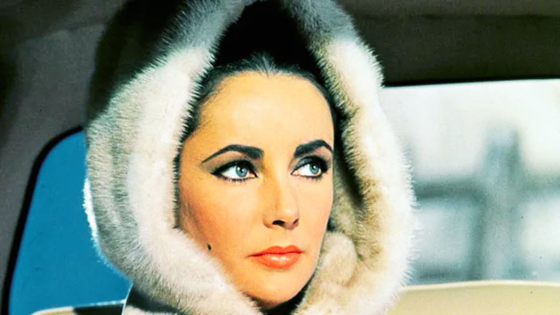 Elizabeth Taylor gazes to the side in a plush white fur hood with bold cat-eye liner and coral lipstick.