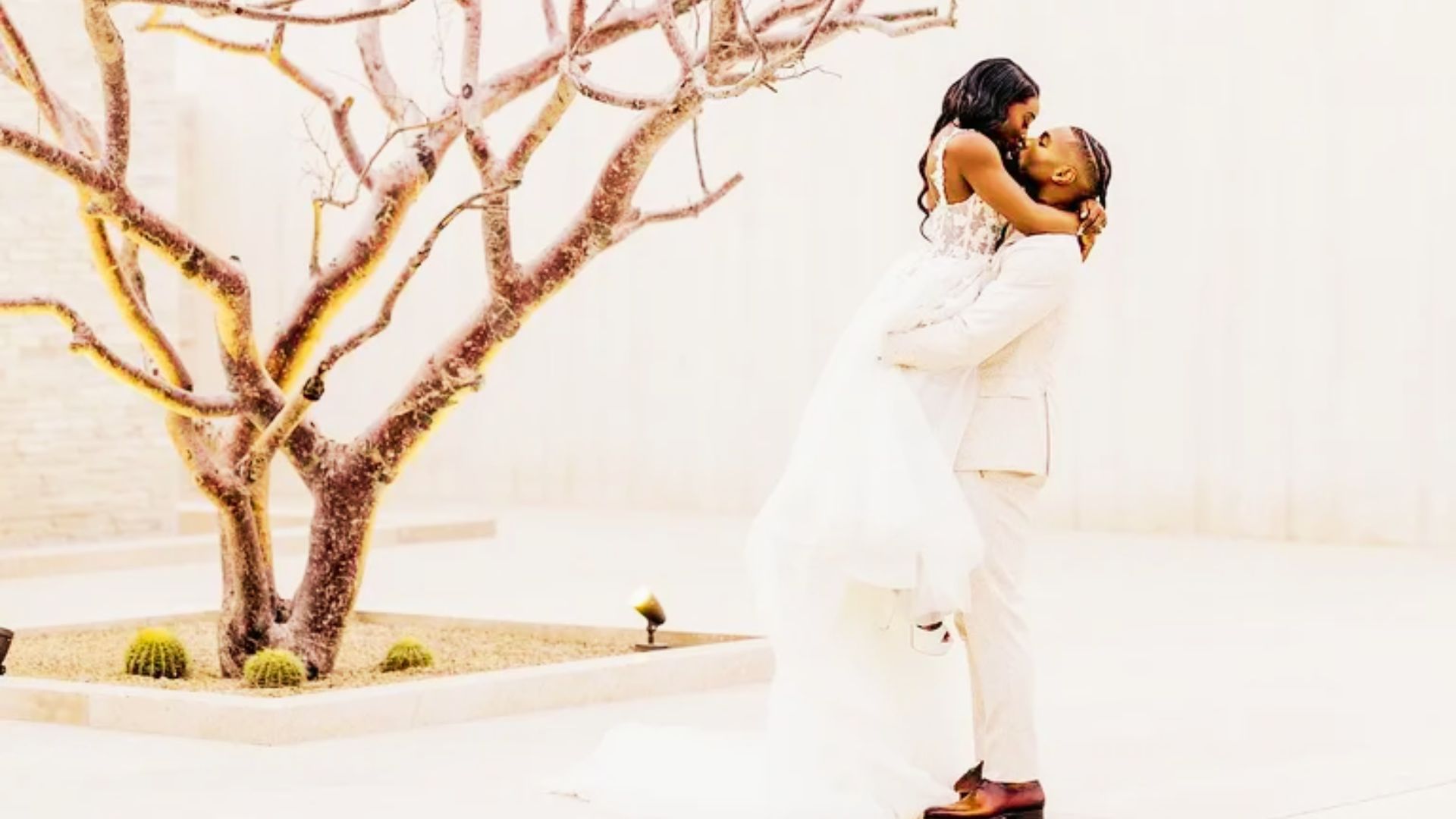 Jonathan Owens lifts Simone Biles as they kiss beside a bare tree and desert landscaping.