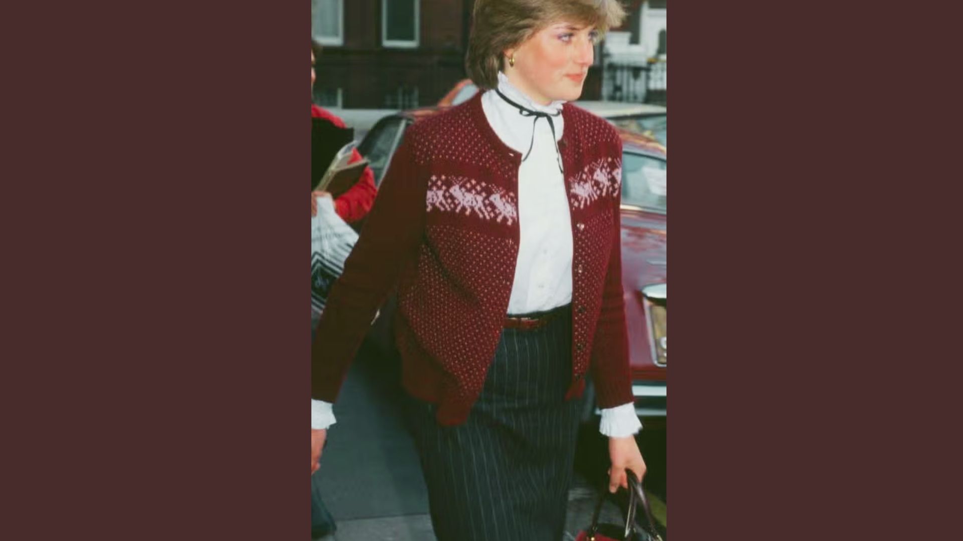 Princess Diana walks outdoors in a burgundy fair-isle cardigan layered over a white ruffled blouse with black ribbon tie.