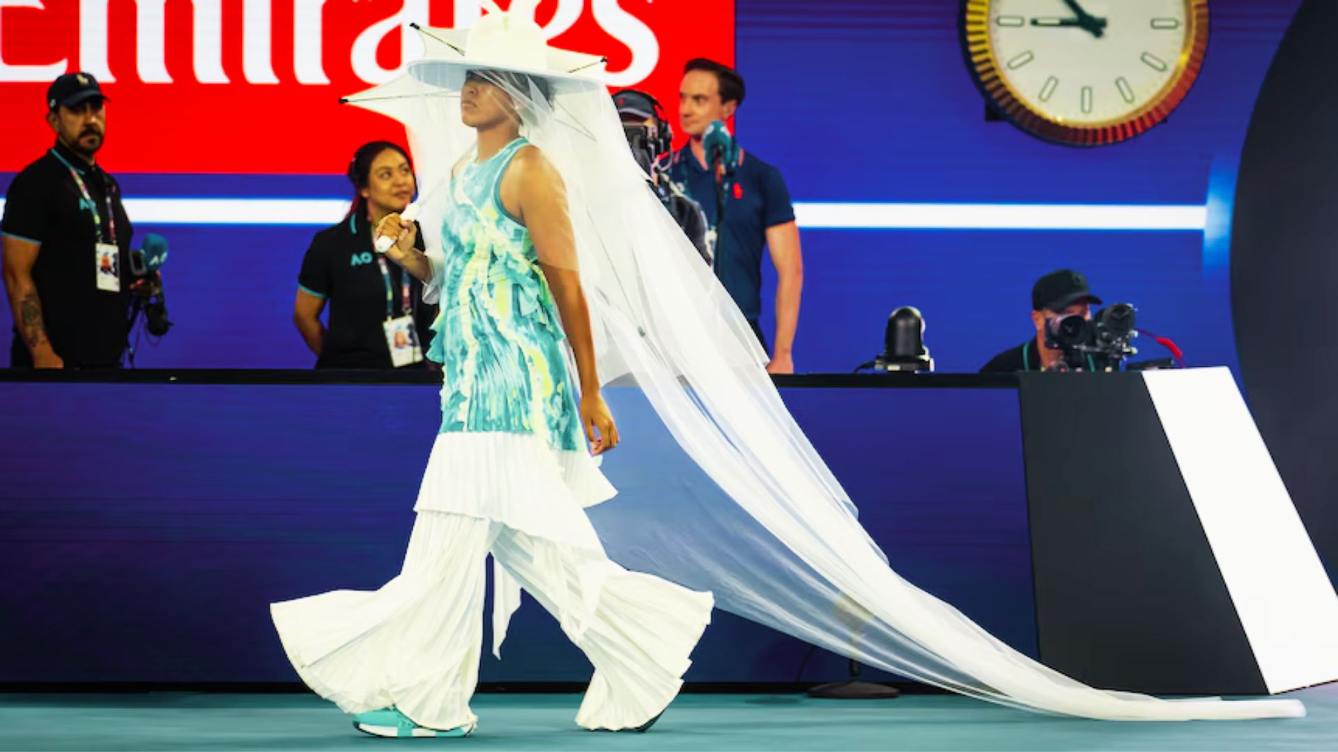 Naomi Osaka makes her Australian Open entrance in a teal tie-dye Nike top, layered over white pleated wide-leg pants.