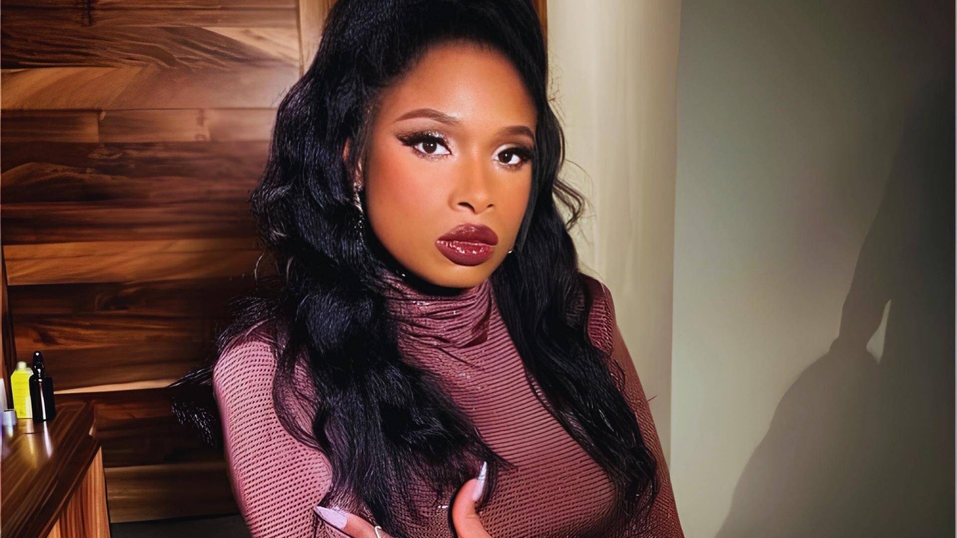 Jennifer Hudson poses in maroon mesh turtleneck with long wavy black hair cascading over shoulders against warm wood panel backdrop.