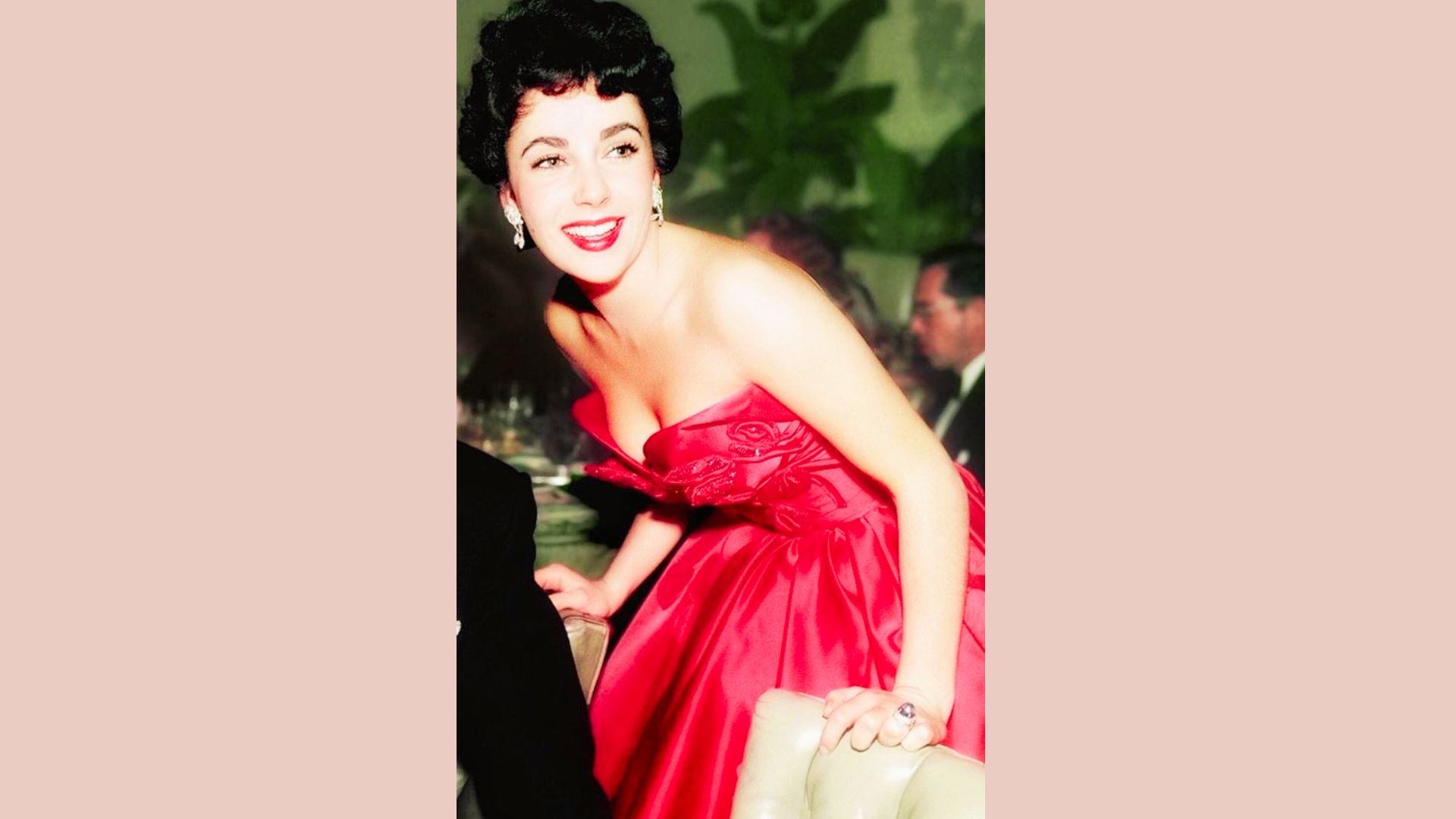 Elizabeth Taylor leans forward in a strapless red satin gown with rose appliqués at a formal event.