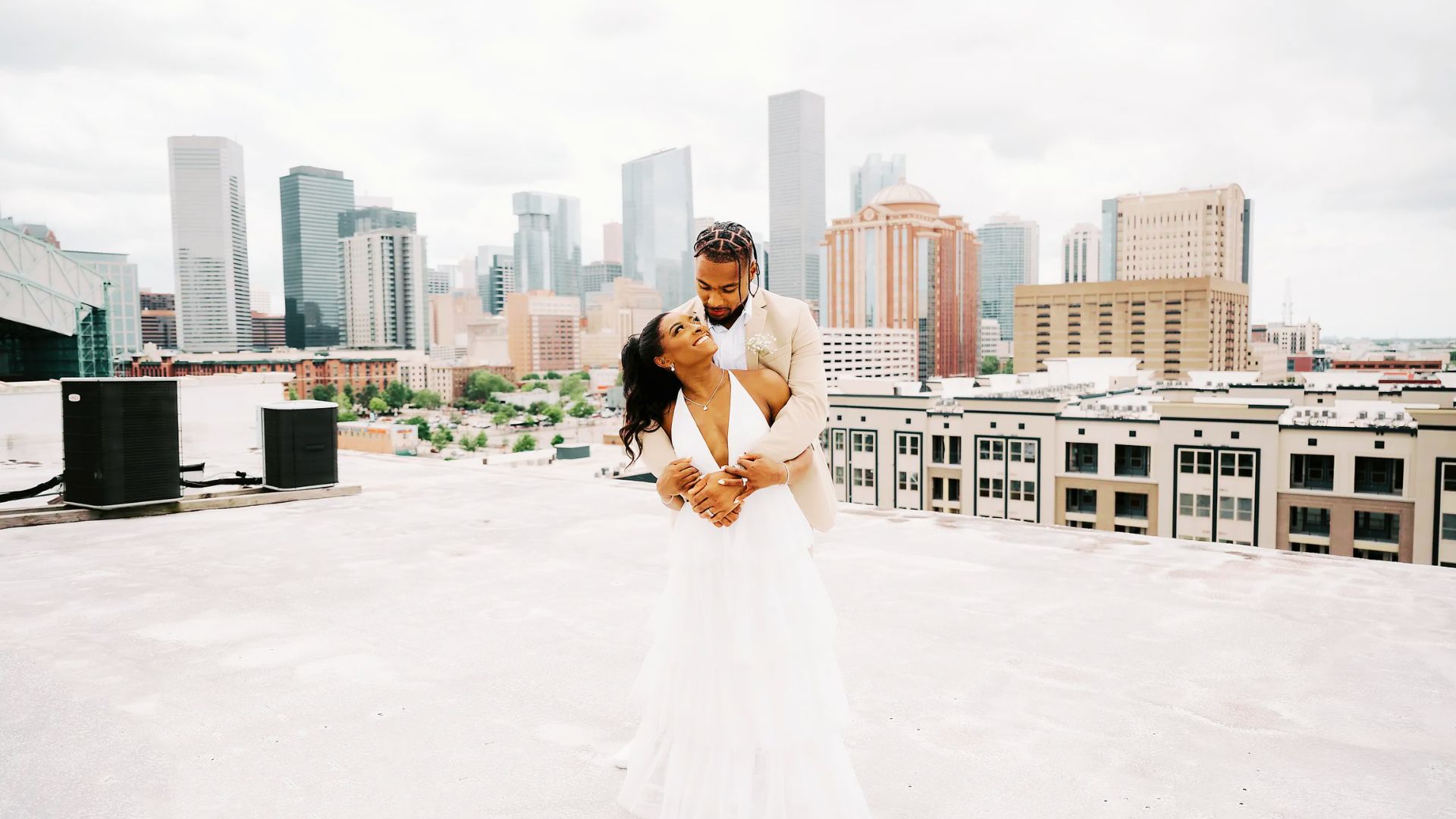 Jonathan Owens embraces Simone Biles from behind on a rooftop with the city skyline behind them.
