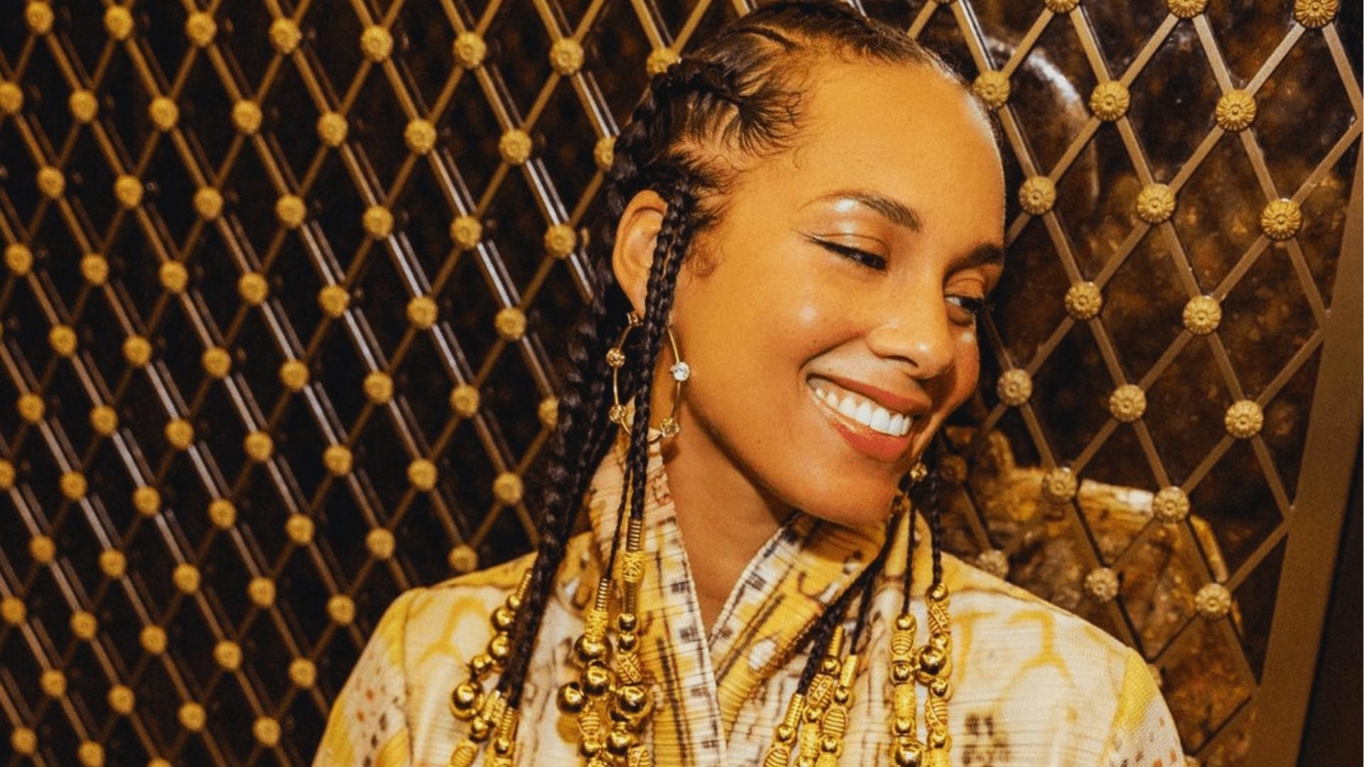Alicia Keys grins in gold patterned jacket with long Fulani braids adorned with gold beads and cuffs against ornate elevator doors.