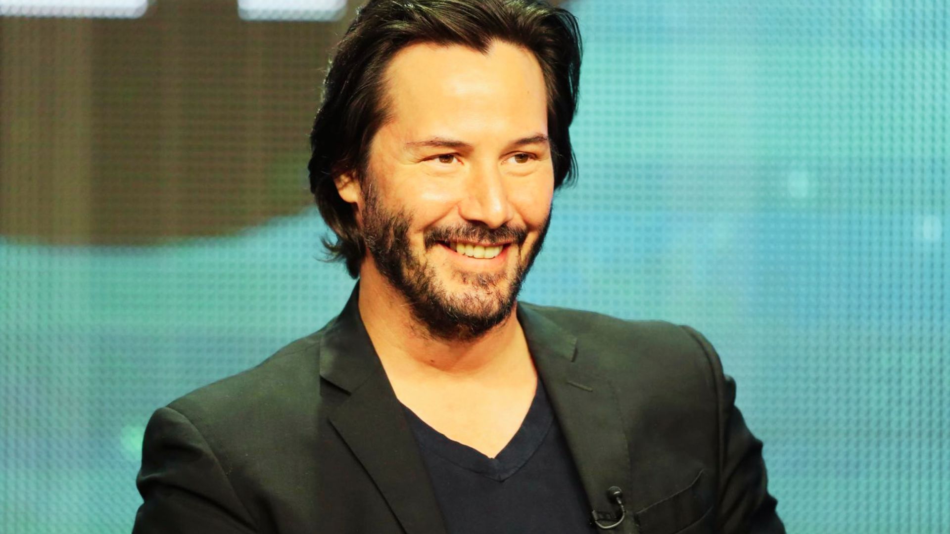 Keanu Reeves in a blazer and black t-shirt smiling.