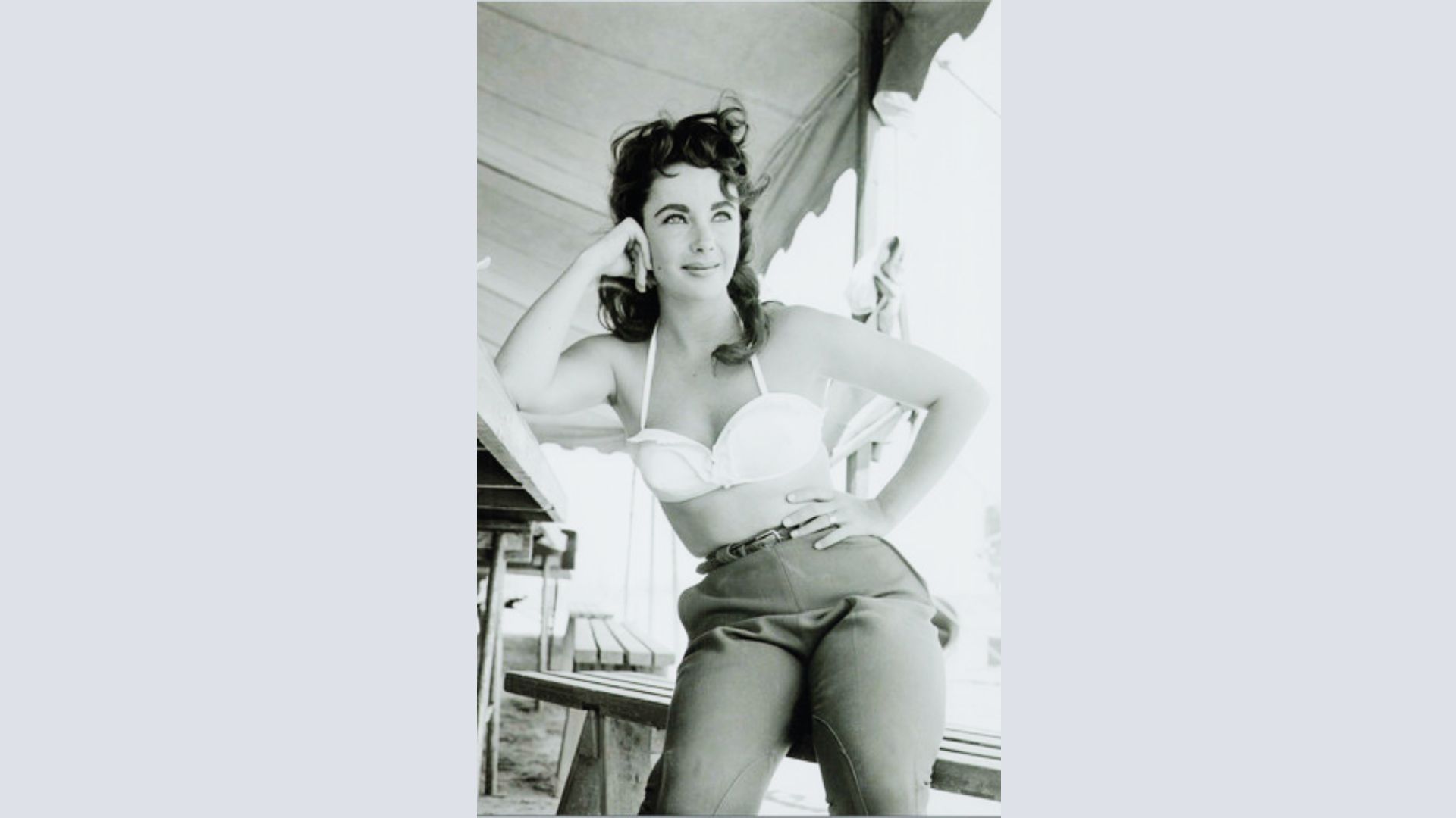 Elizabeth Taylor relaxes outdoors in a white halter bikini top and high-waisted dark pants.