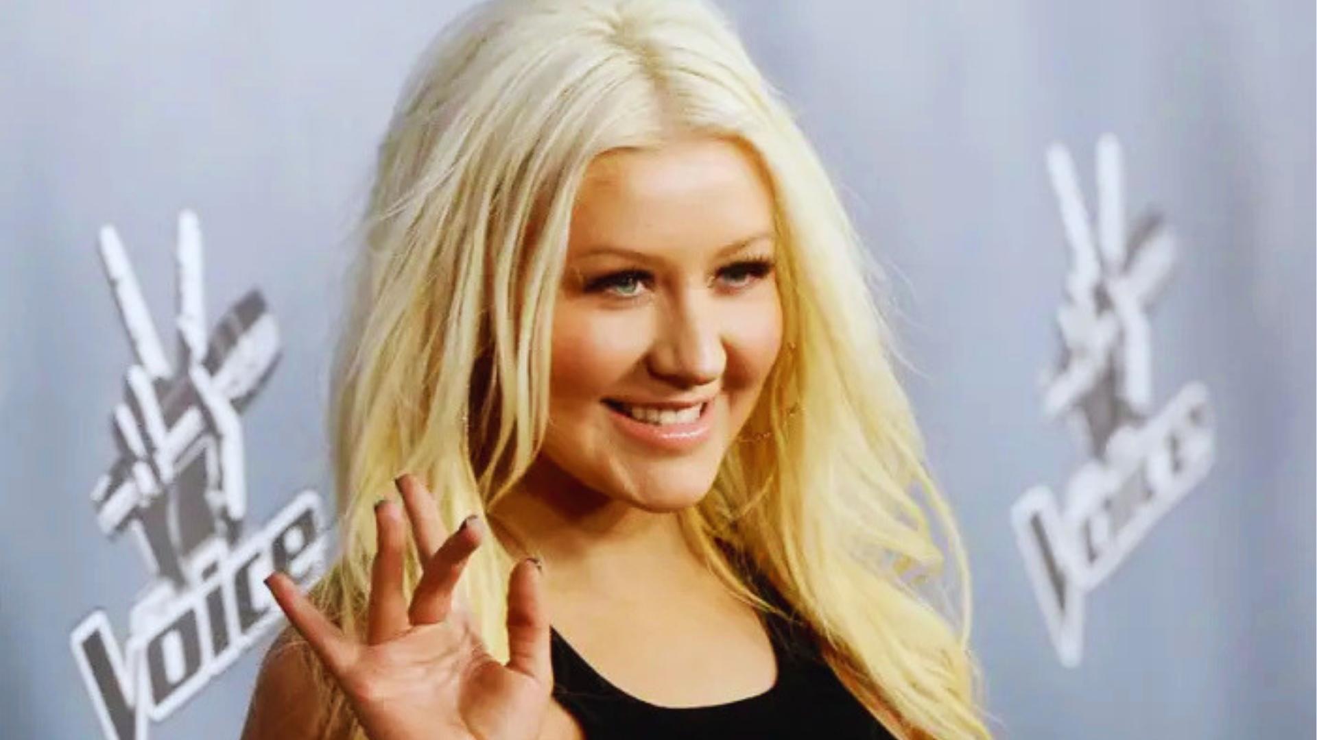 Christina Aguilera smiles at The Voice backdrop with platinum blonde hair, light mascara, and natural glowy skin in black top.