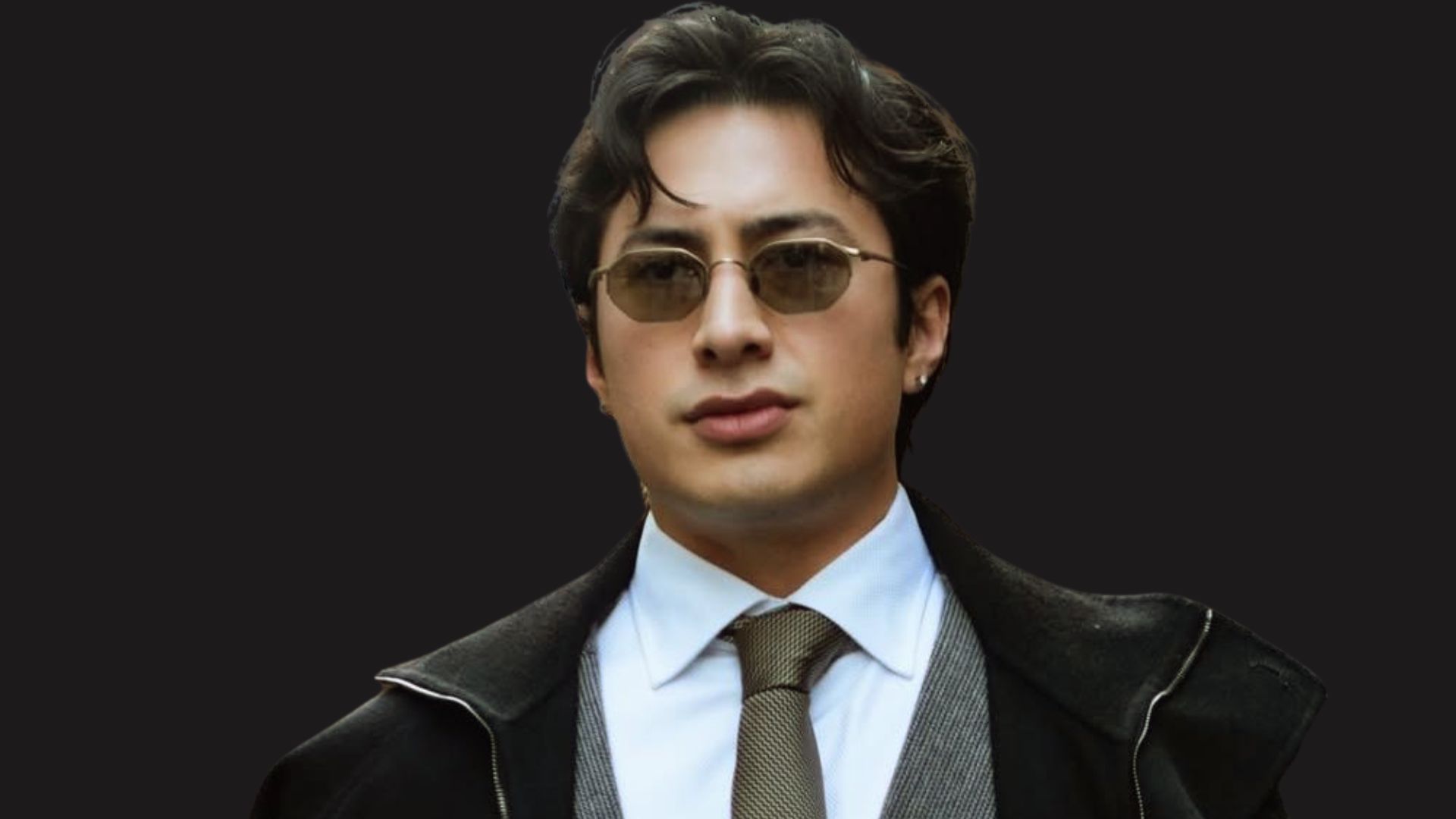 Hudson Williams with voluminous dark brown hair wearing tinted sunglasses, a tie, and a black coat against a dark background.