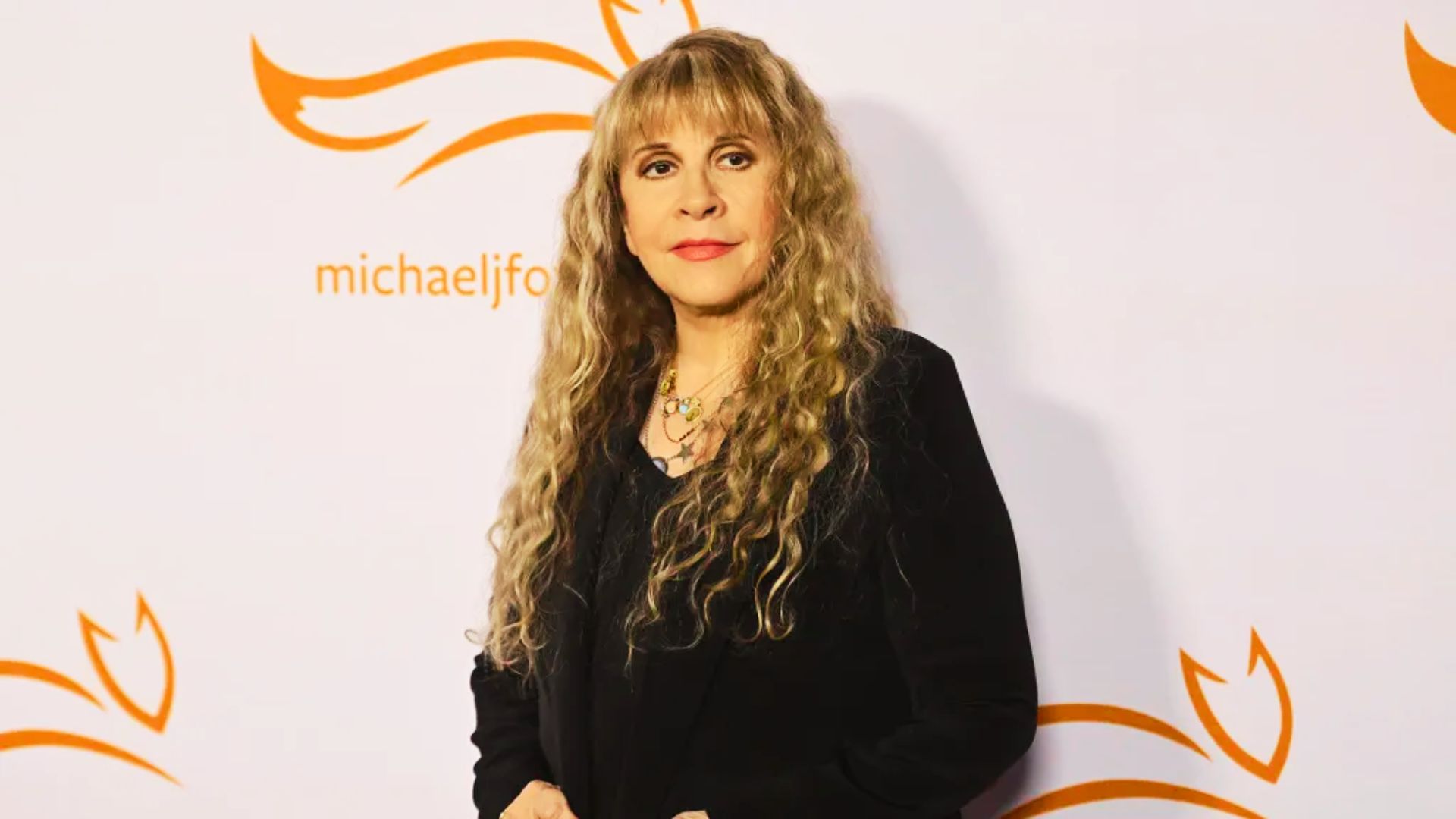 Stevie Nicks stands at an event wearing black, with long blonde curly hair and wispy bangs.