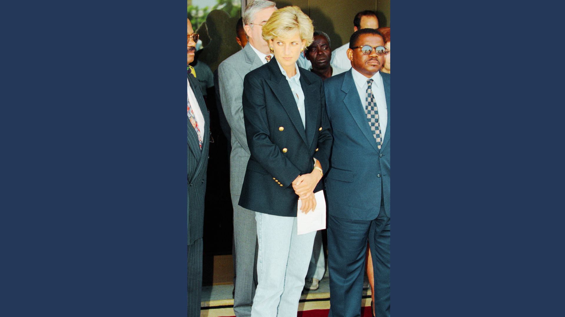 Princess Diana stands among a group wearing a navy double-breasted blazer with gold buttons over a light blue shirt and pale jeans.