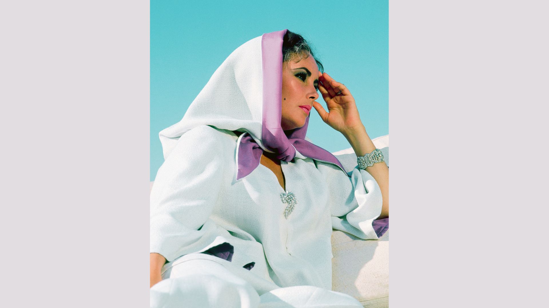 Elizabeth Taylor shields her eyes in a white hooded caftan with lavender silk trim against a bright blue sky.