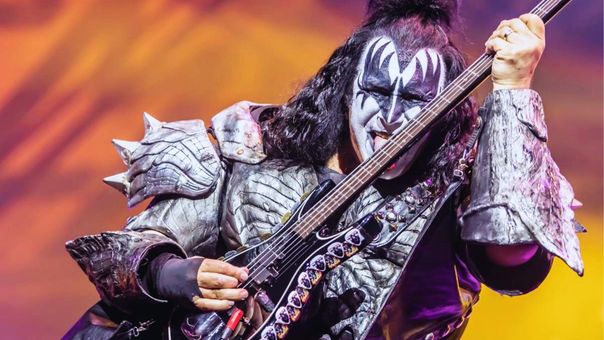 Gene Simmons performs as The Demon with full white and black face paint, spiked armor, tongue out, holding bass onstage.