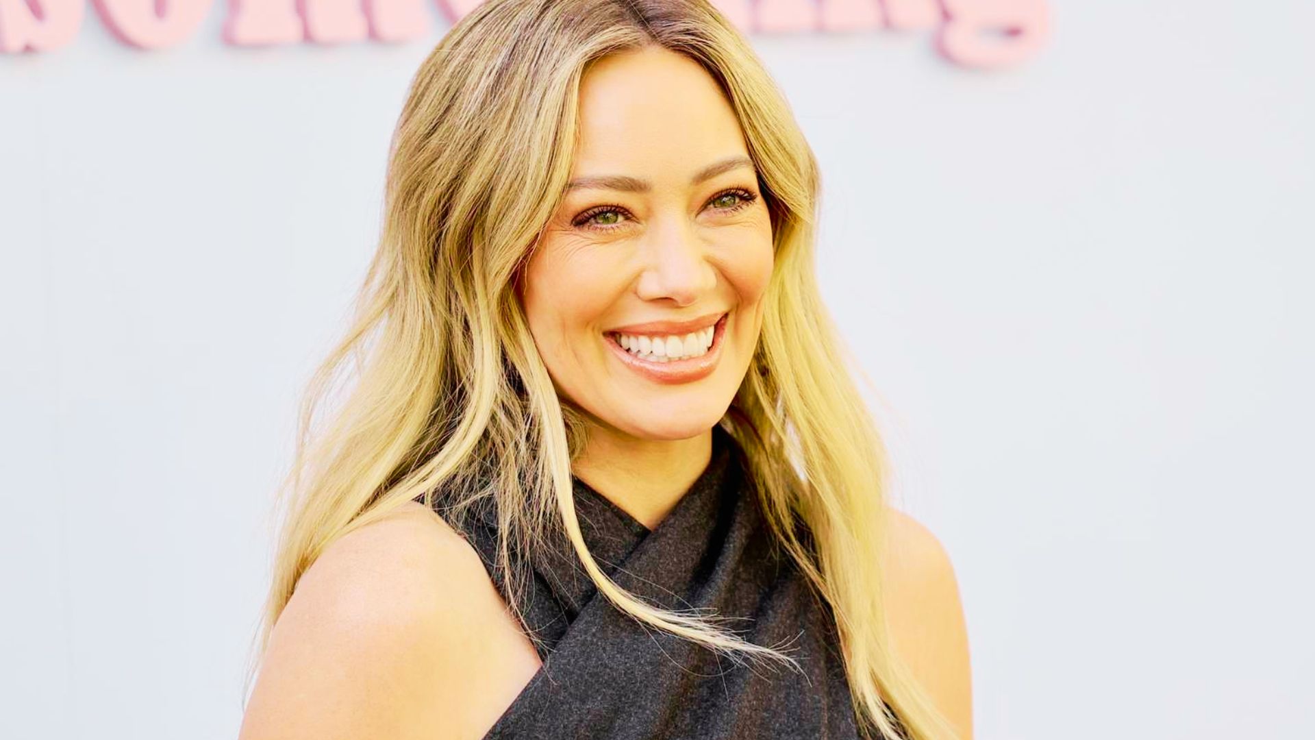 Hilary Duff smiles at a press event in a dark gray sleeveless crisscross halter top.