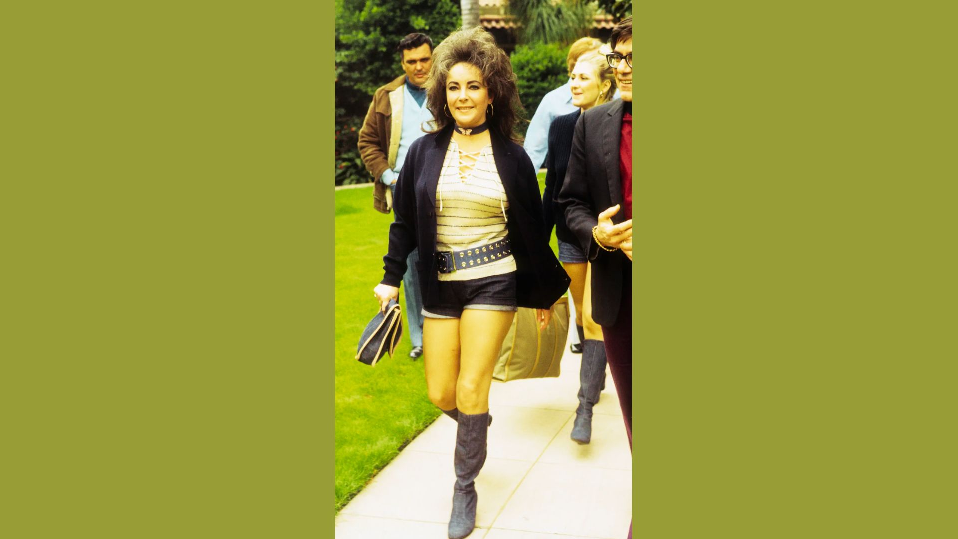Elizabeth Taylor walks outdoors in a 1970s ensemble lace-up striped top, dark micro shorts, wide studded belt, and suede knee-high boots.