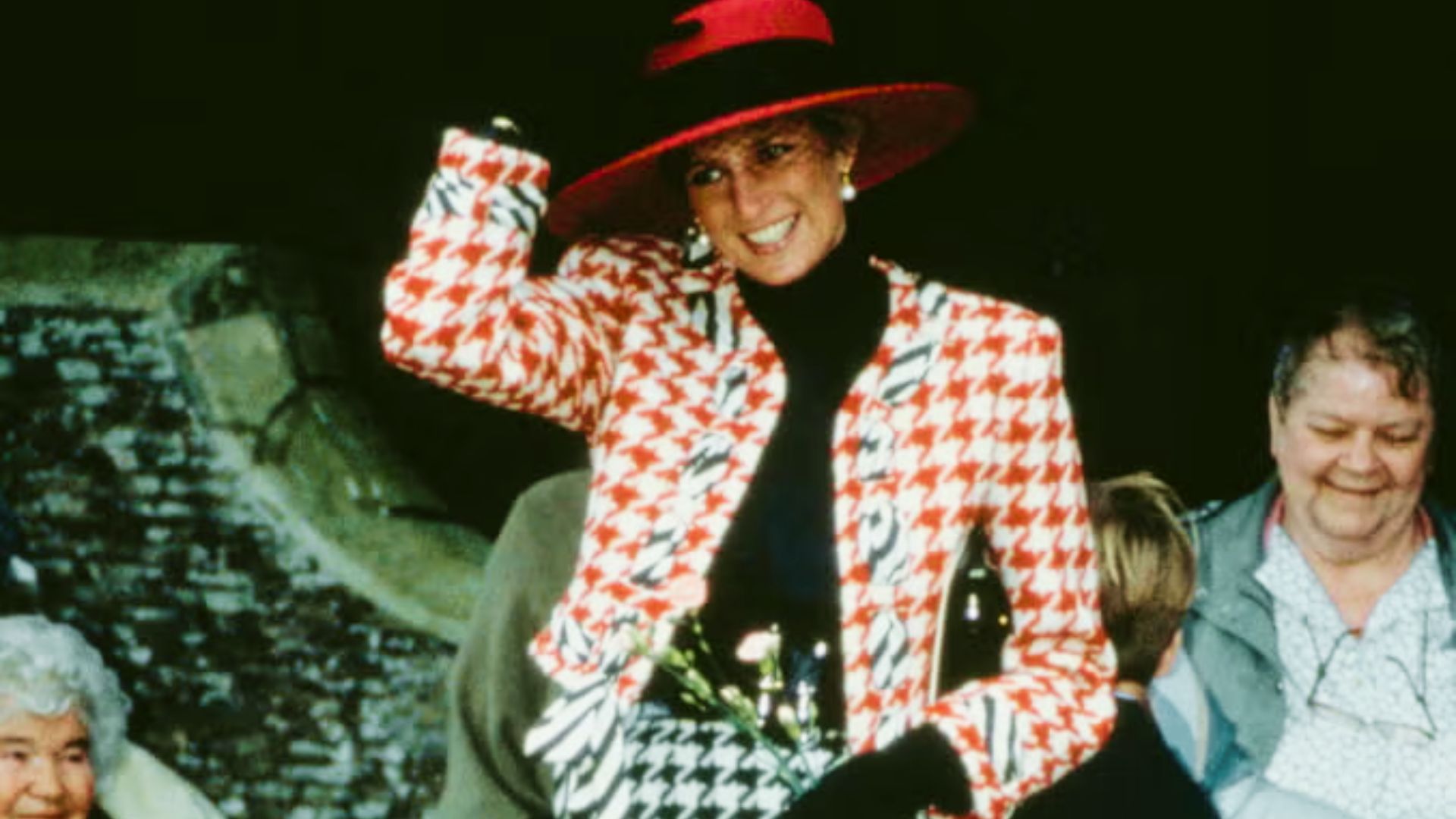 Princess Diana waves to a crowd in a red and white houndstooth blazer layered over a black turtleneck.